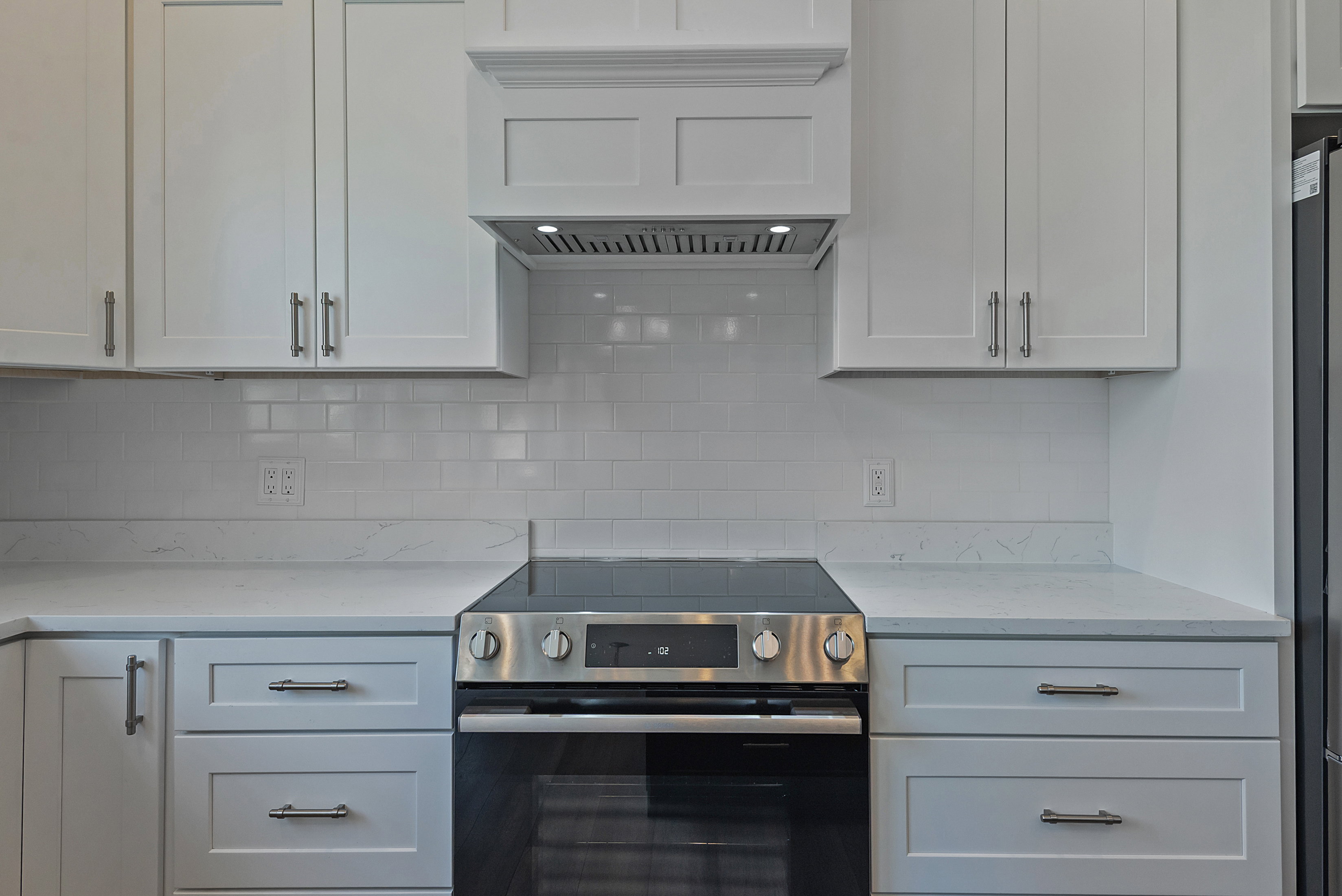 Kitchen featuring white shaker-style cabinetry, a built-in stainless steel electric range, a matching range hood with integrated lighting, a subway tile backsplash, and light-toned countertops