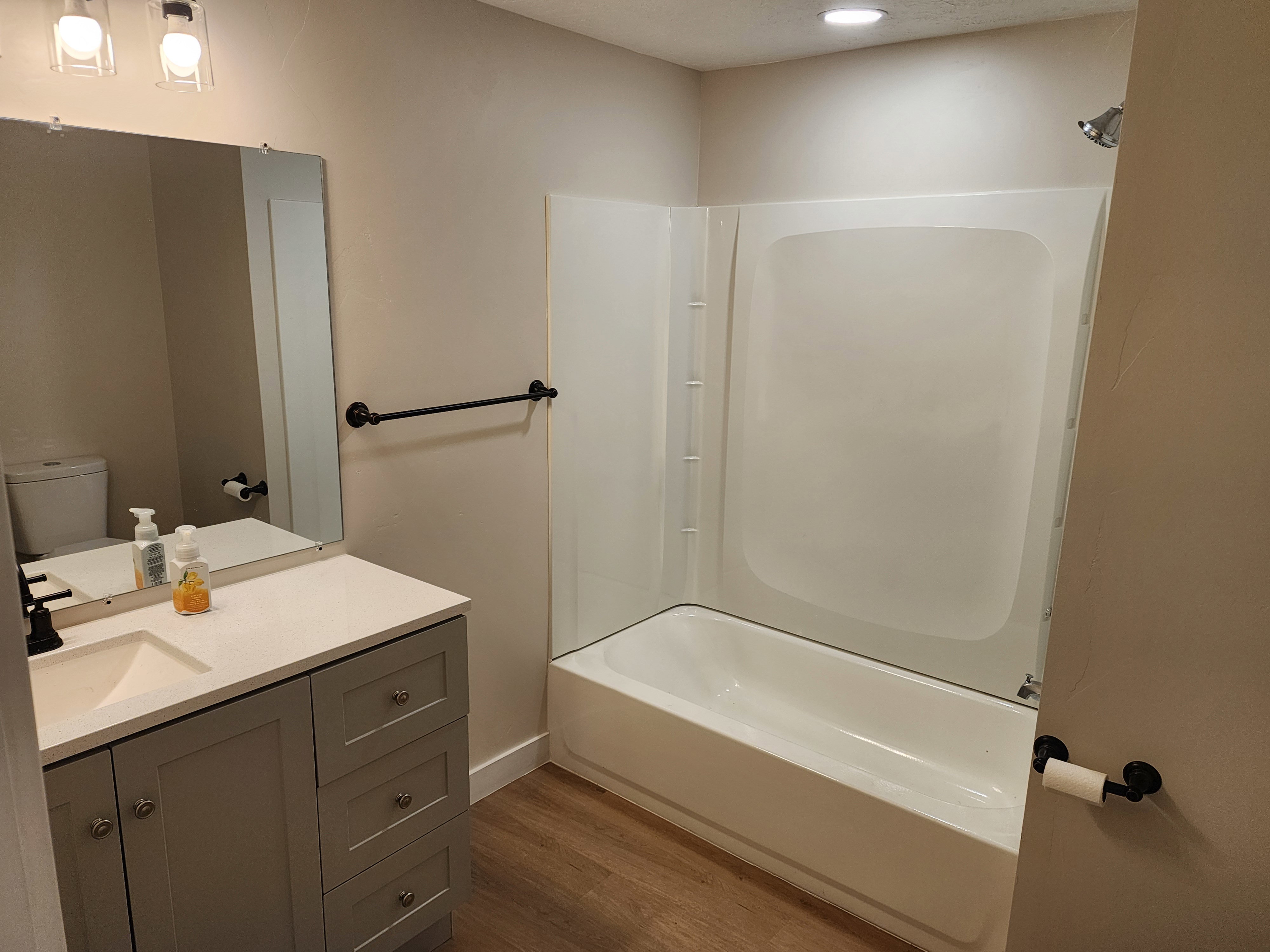 Bathroom featuring a white tub and shower unit, a light gray vanity with a single sink and matte black hardware, a rectangular mirror, and wood-finish flooring