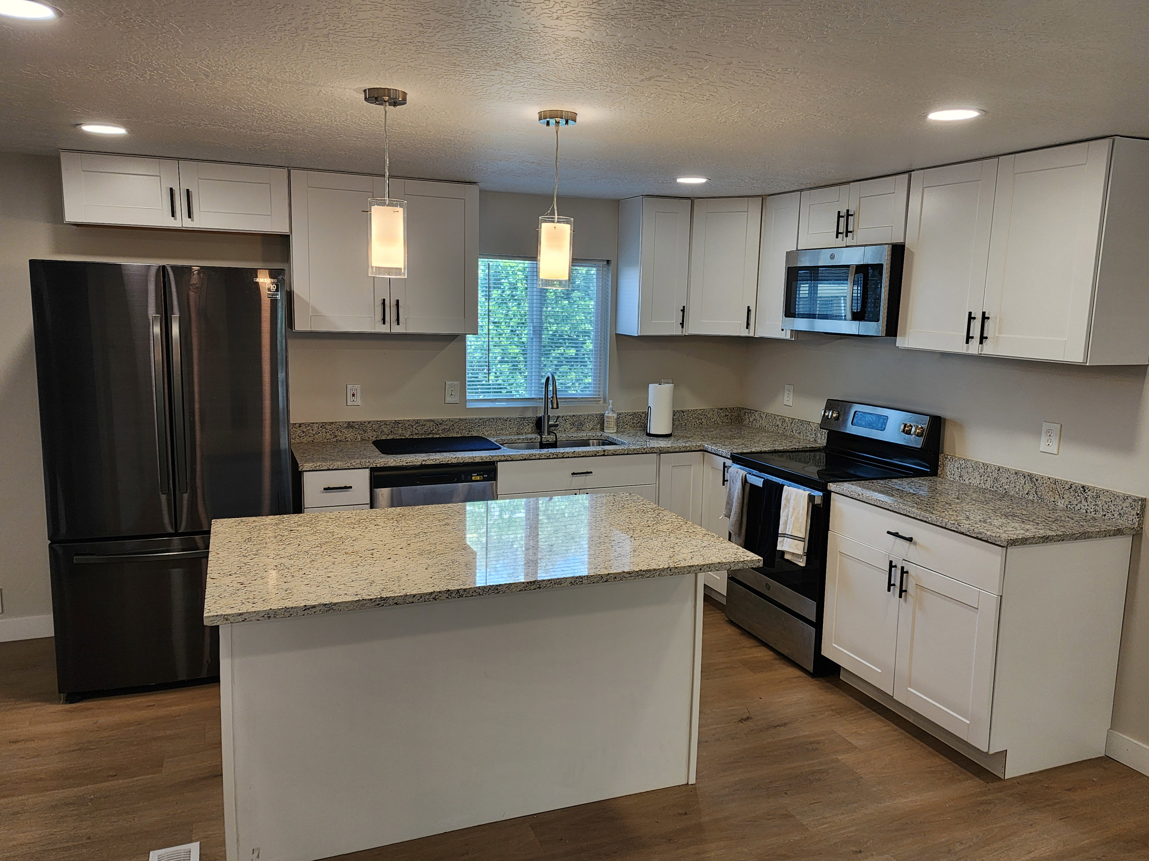 Modern kitchen with white shaker cabinetry, speckled countertops, stainless steel appliances, a central island, and wood-finish flooring