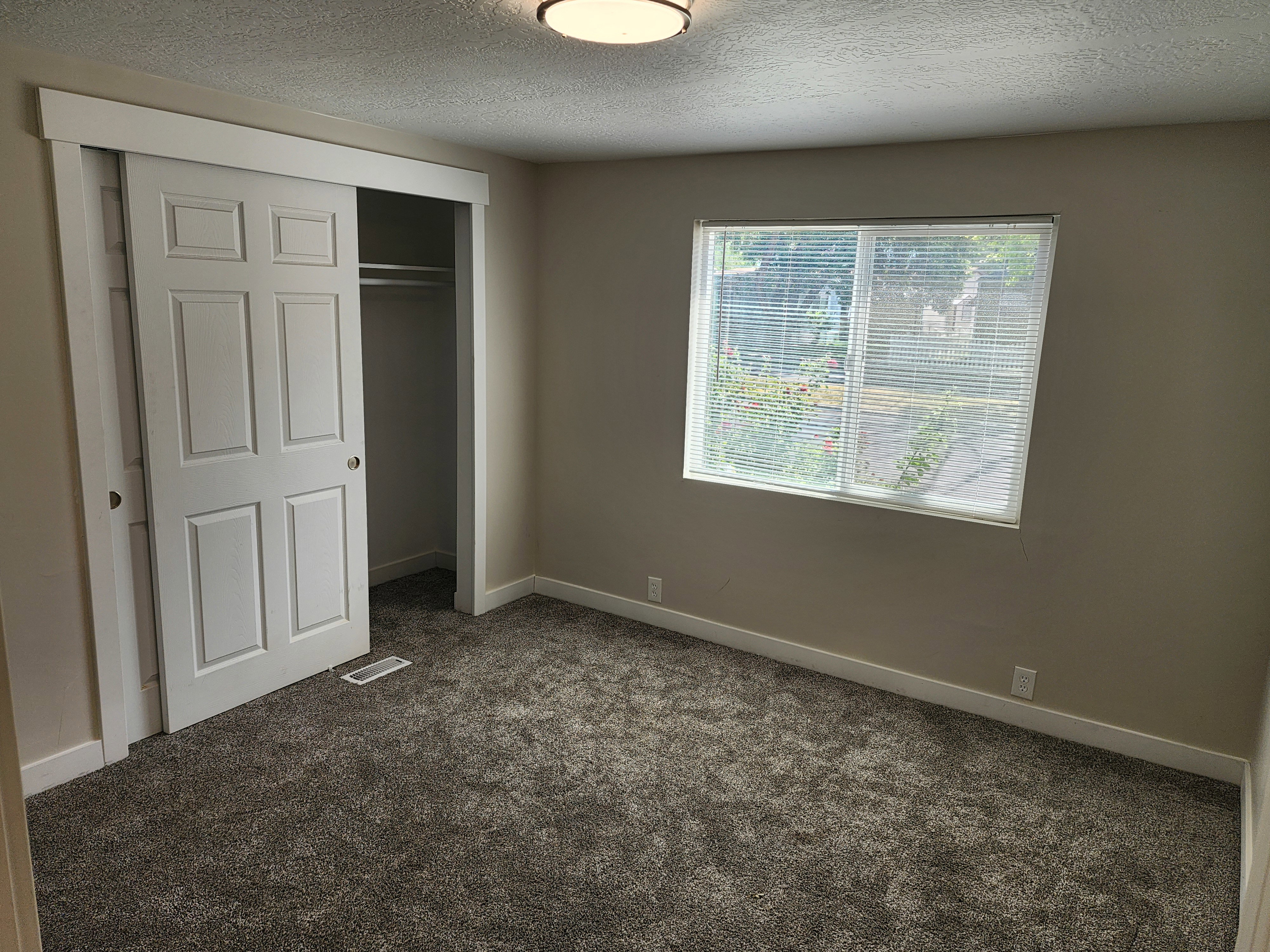 Carpeted room featuring a window with blinds, a ceiling-mounted light fixture, sliding panel closet doors, and white baseboards