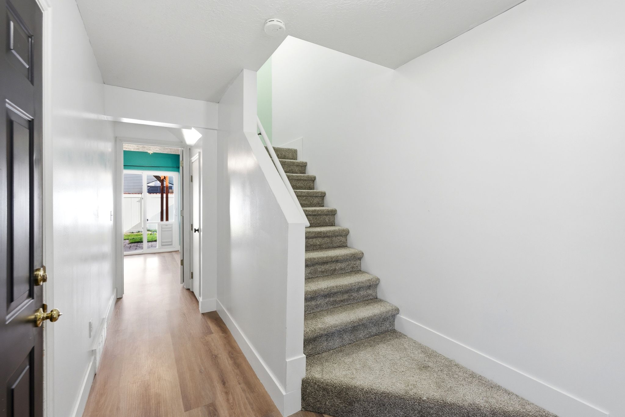Entryway featuring wood-finish flooring extending into the hallway, a carpeted staircase with a white railing, a dark paneled front door with brass hardware, and a distant view of a backyard through glass doors