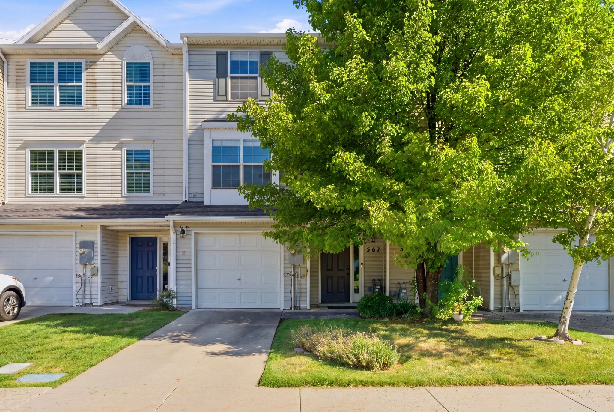 Multi-story townhome featuring light-colored siding, a prominent front entry with a blue door, an integrated garage, and a lush green lawn