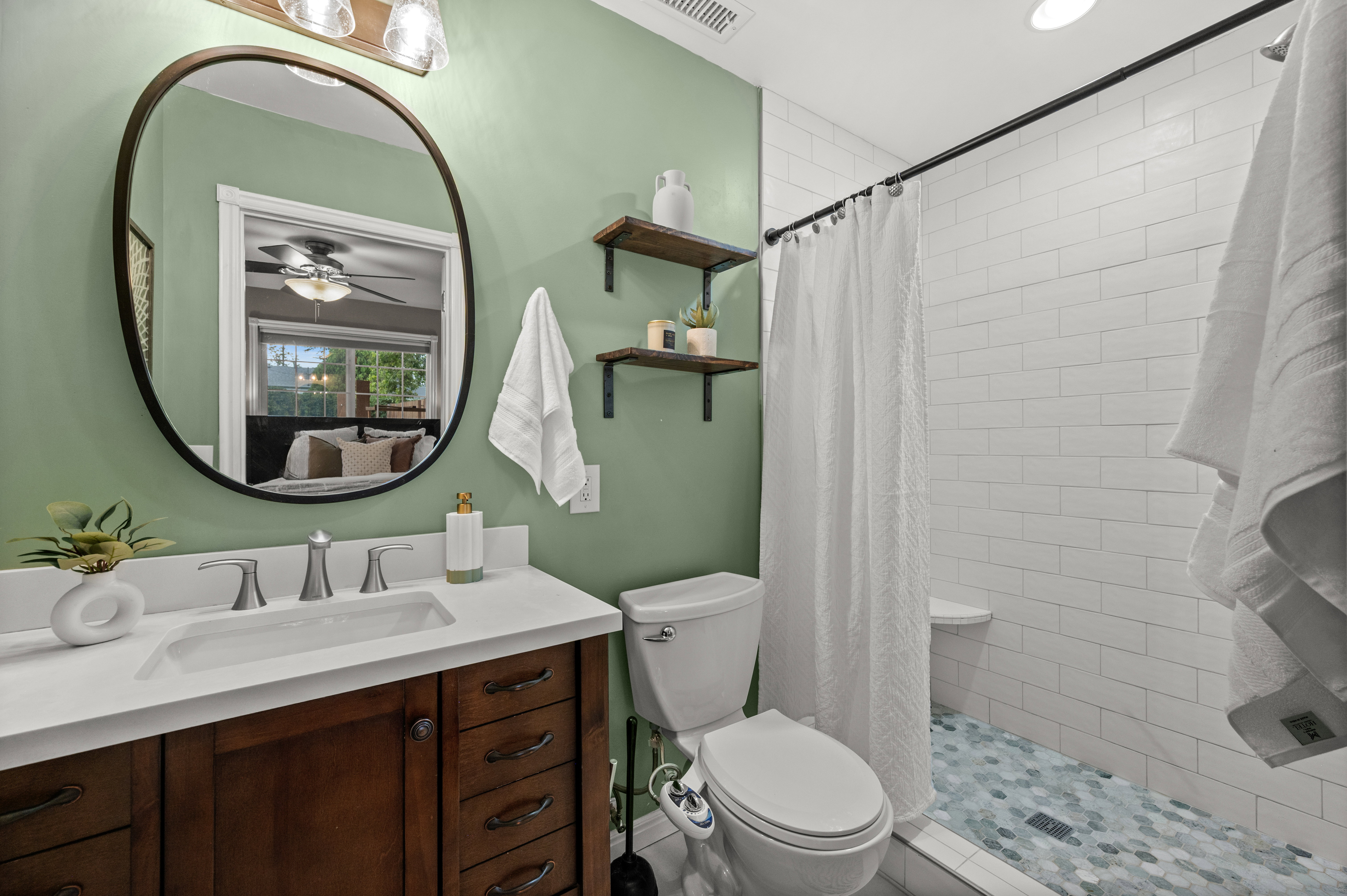 Bathroom featuring a dark wood-finish vanity with a white countertop and rectangular undermount sink, an oval mirror, and a tiled shower with a built-in bench and hexagonal floor tiles