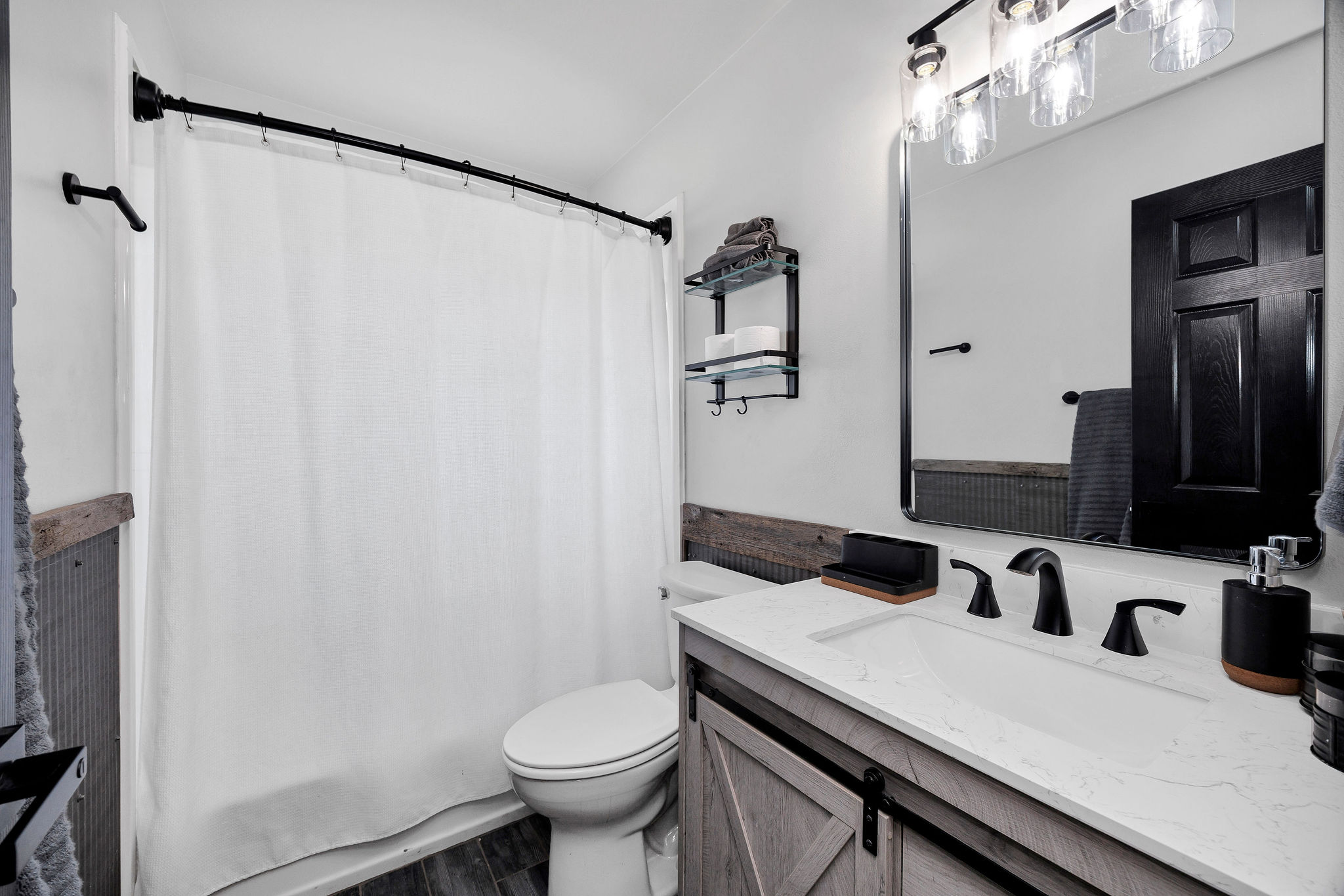 Bathroom featuring a shower-tub combination with a curved rod, a vanity with a barn door style cabinet, a white countertop with an integrated sink, a rectangular framed mirror, and a three-light vanity fixture