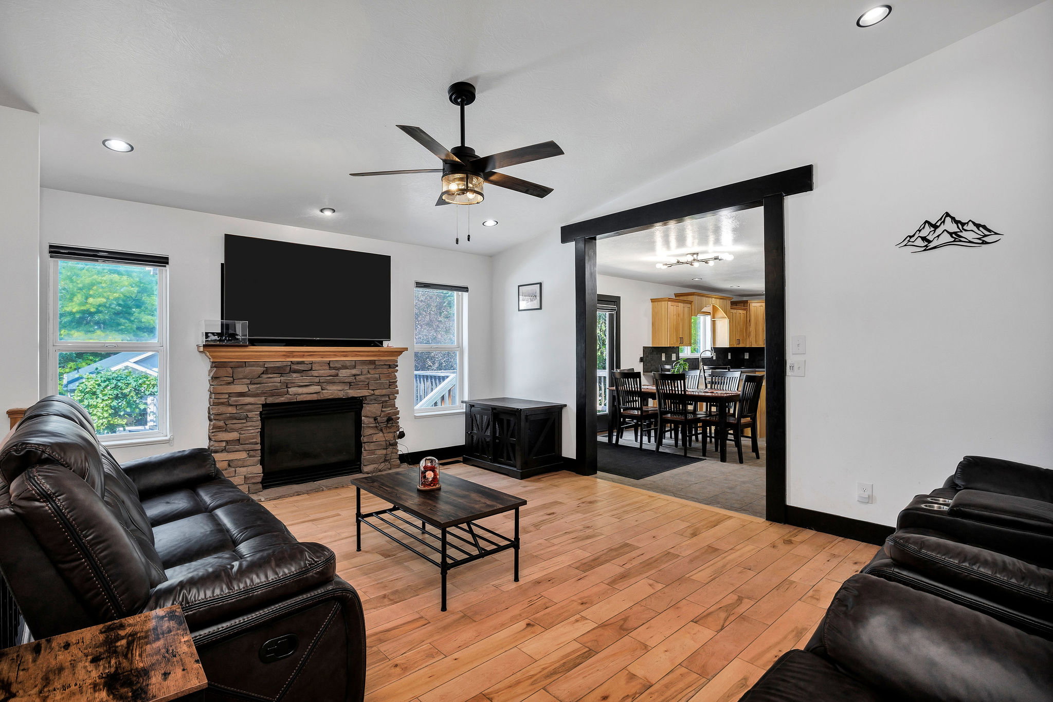 Living area featuring a stone-clad fireplace with wood mantle, light wood-finish flooring, a ceiling fan with integrated lighting, recessed lighting, and an expansive doorway opening to an additional room