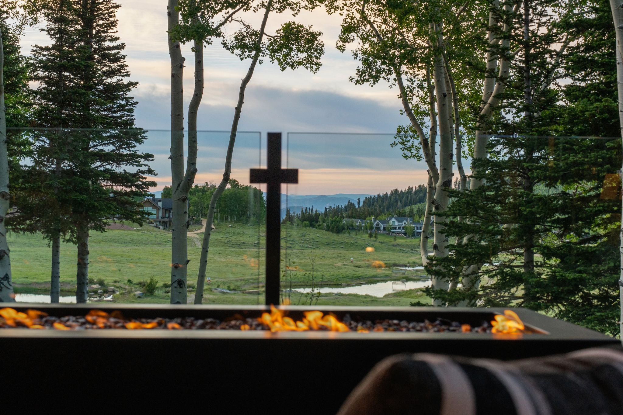 Outdoor fire feature with a black metal frame, glass panel railing, and surrounding evergreen and aspen trees