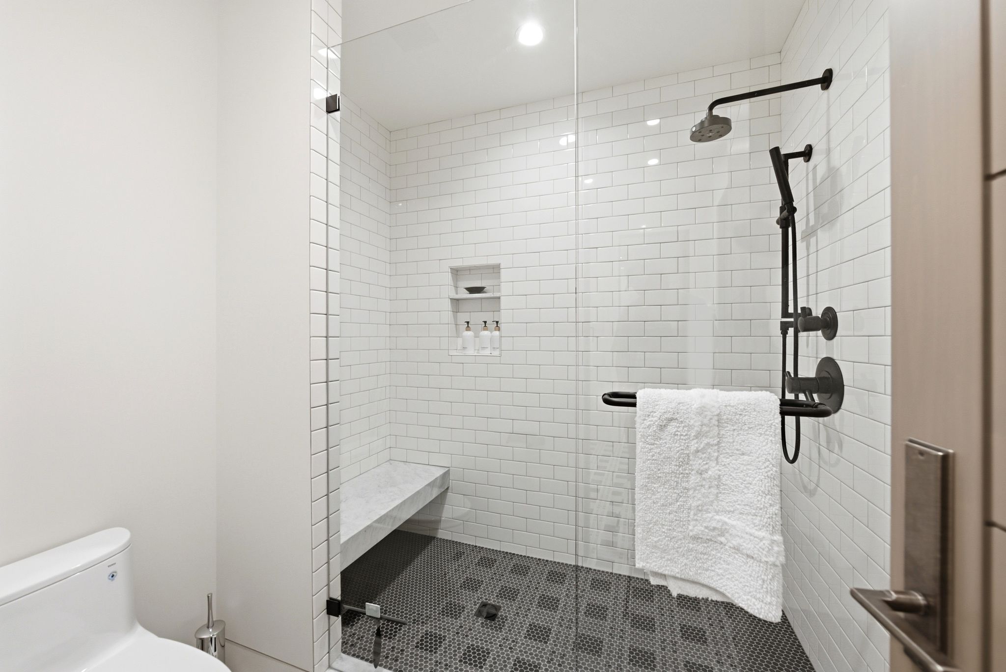 Frameless glass shower enclosure featuring matte black fixtures, subway tile walls, a built-in bench, and hexagon tile flooring