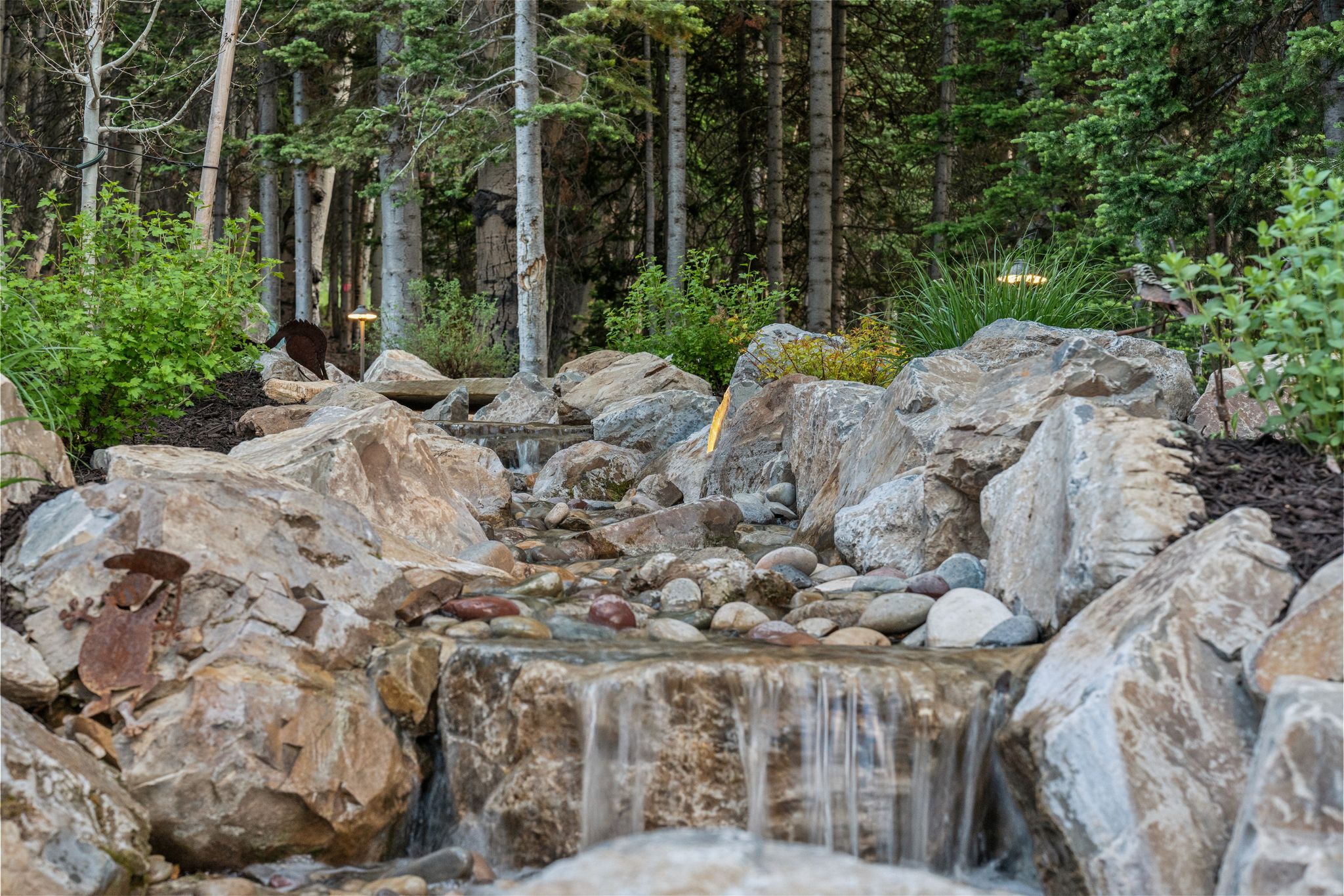 Natural stone water feature with integrated lighting, surrounded by mature trees and varied foliage