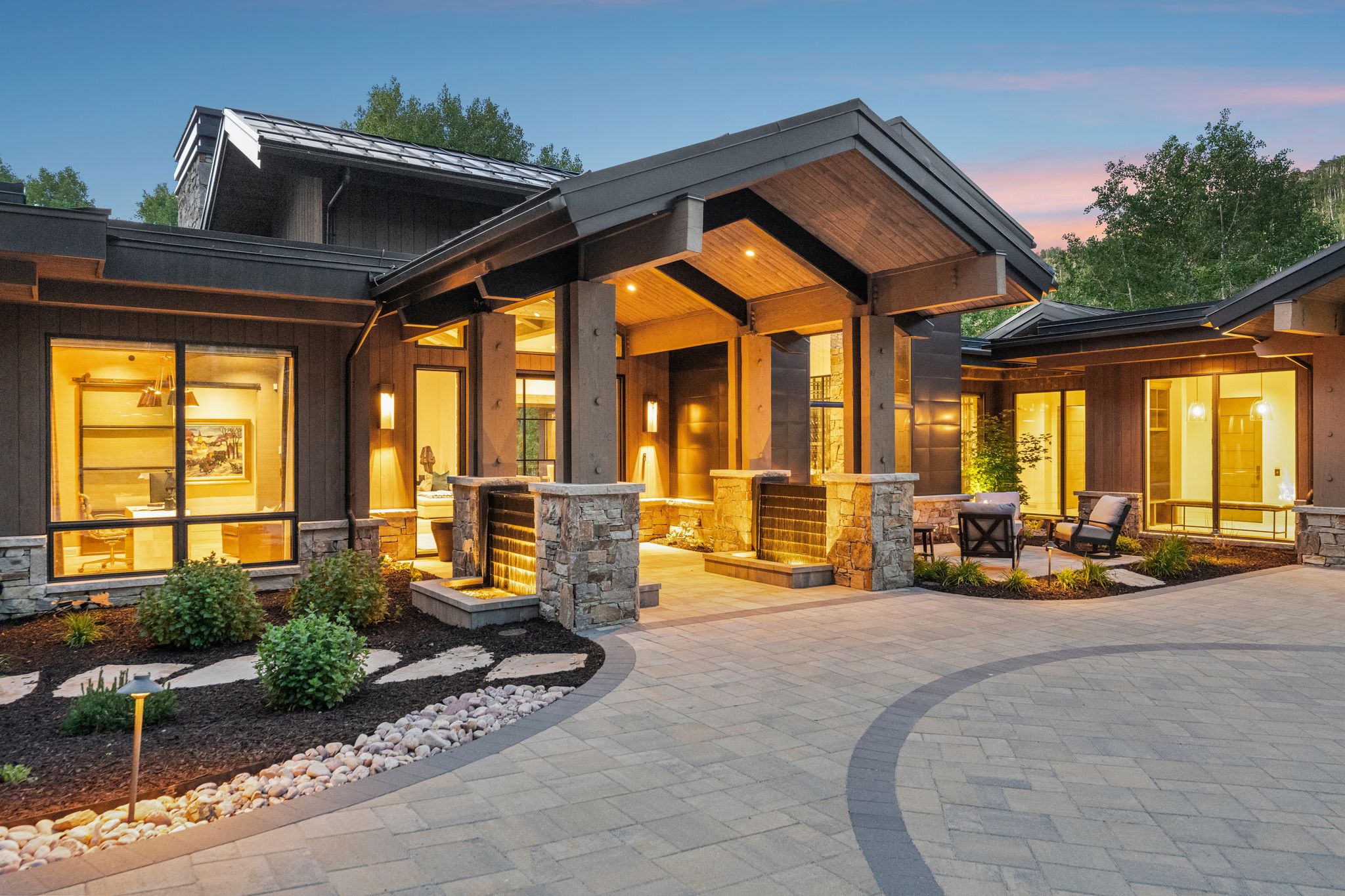 Contemporary exterior featuring a stone paver driveway, an inviting entry patio, a stone veneer water feature, a wood-paneled ceiling on the portico, and expansive windows