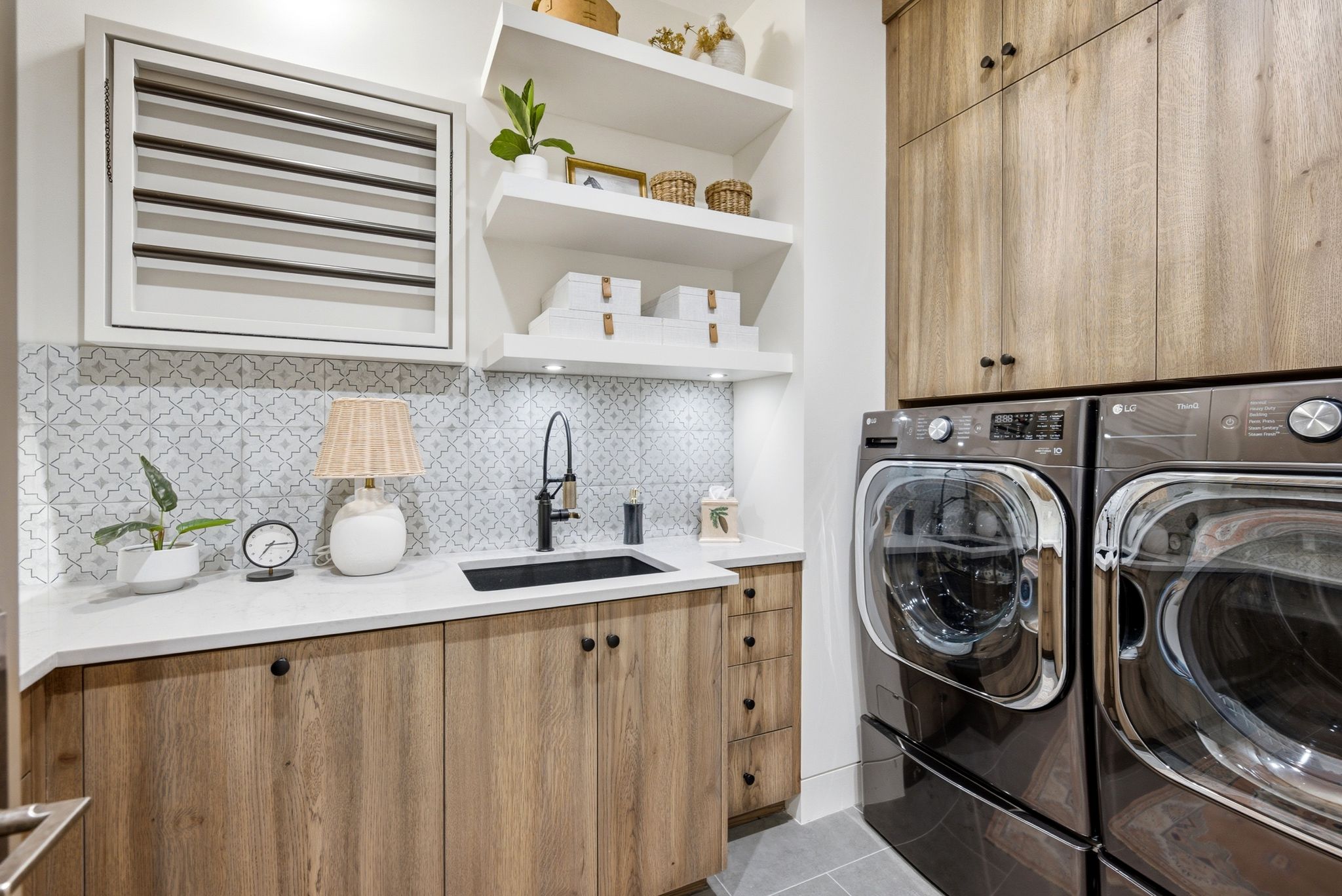 Laundry area featuring wood-finish cabinetry, a white countertop, patterned tile backsplash, and a black undermount sink with a matte black faucet