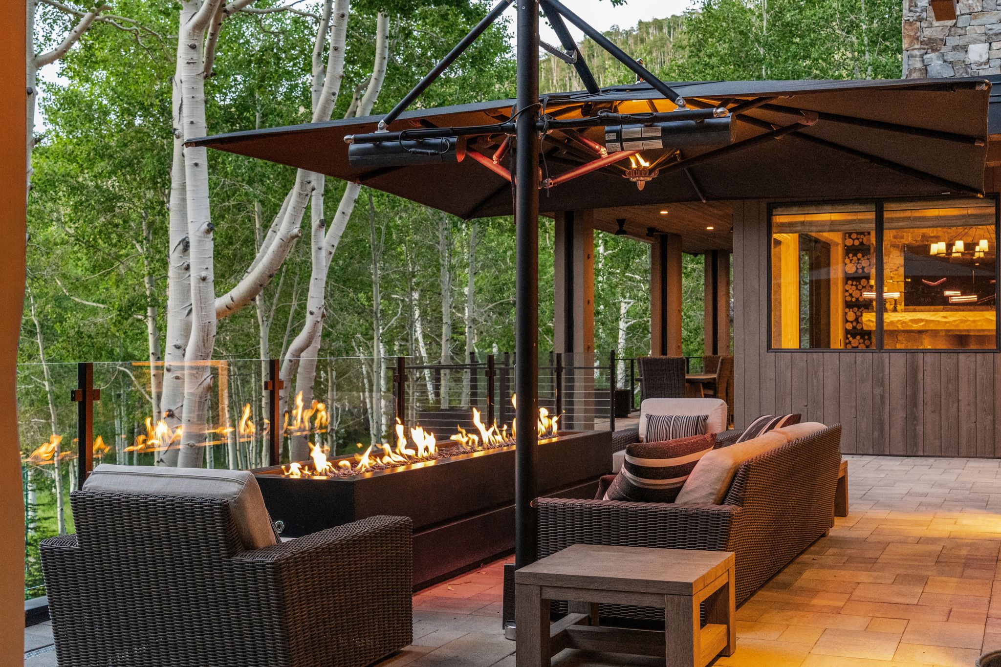 Expansive outdoor patio featuring a linear fire pit with glass barrier, paver flooring, a large cantilever umbrella, and a glass railing system