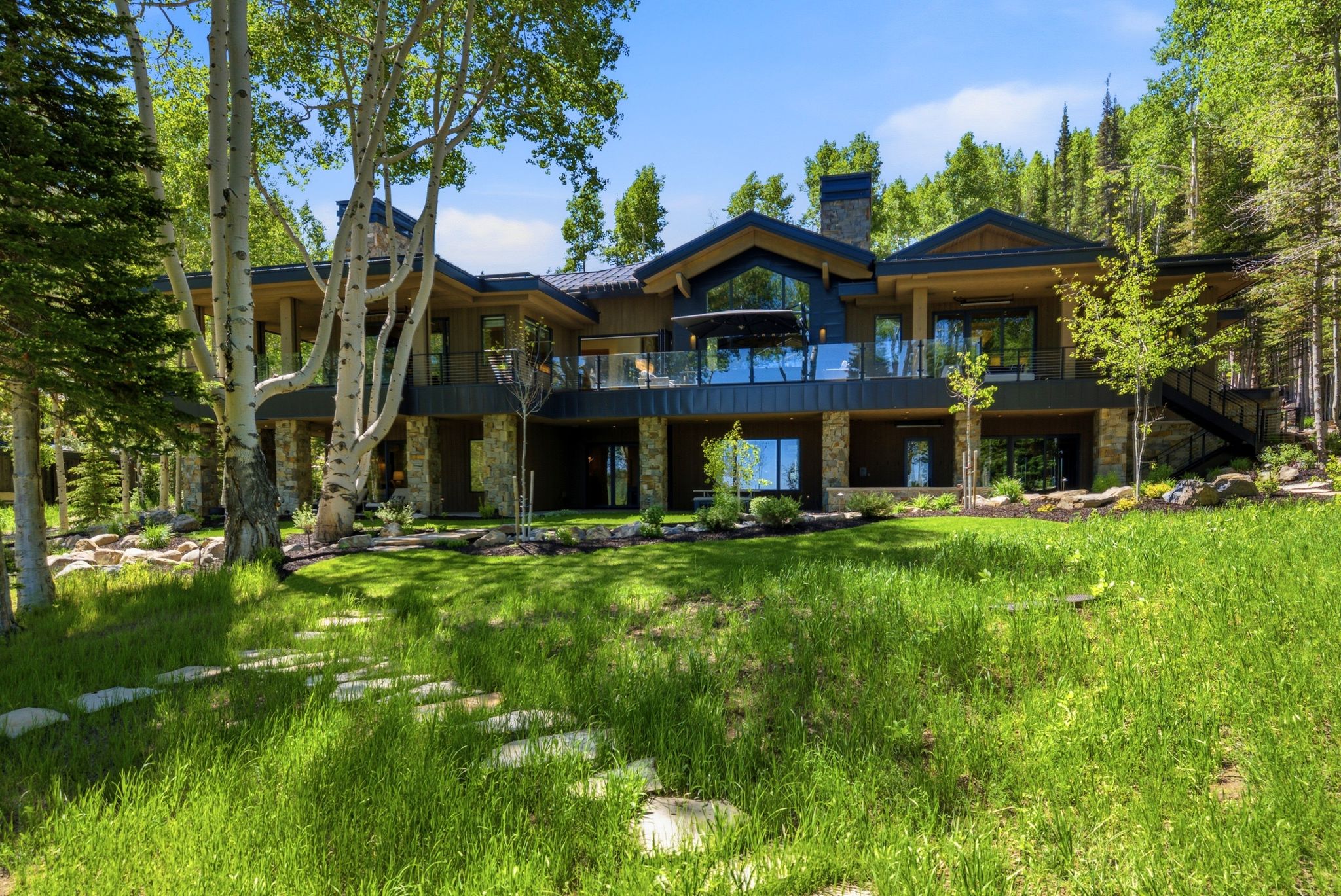 Contemporary mountain residence featuring multiple balconies with glass railings, a stone chimney, and an expansive lawn