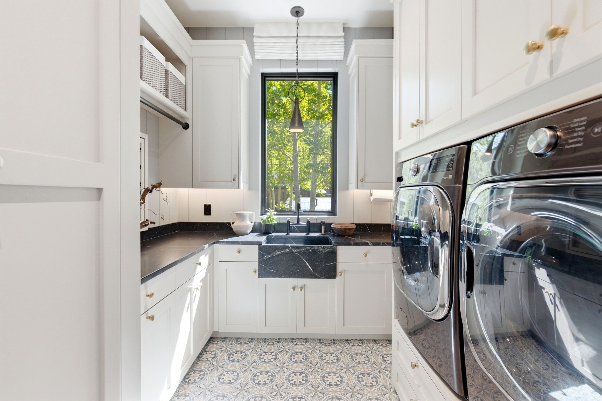 White shaker cabinetry with gold-toned hardware, black solid surface countertops, and patterned floor tiles define this utility space