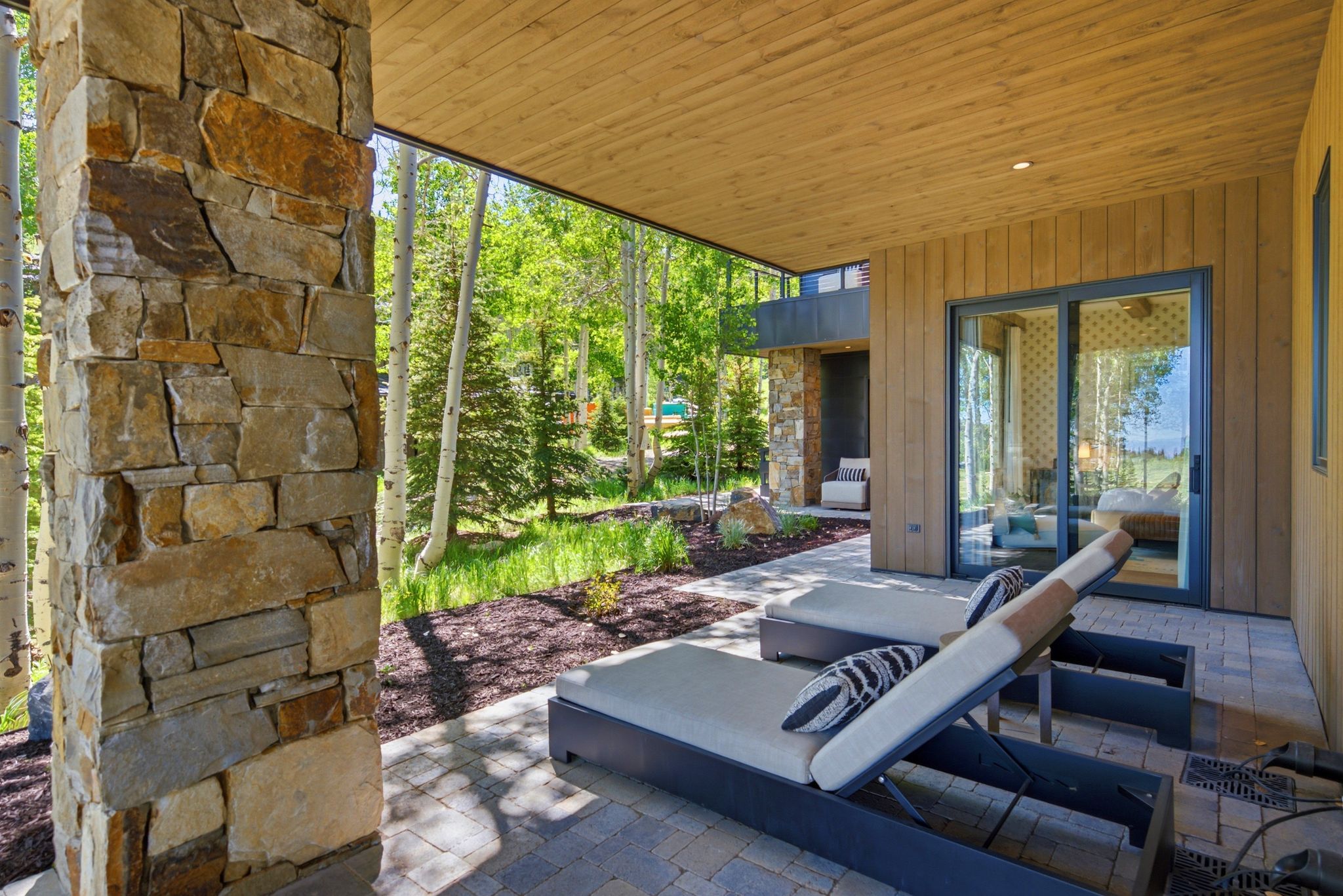 Expansive stone paver patio featuring a natural wood-finish ceiling, a stone-clad column, and vertical wood siding