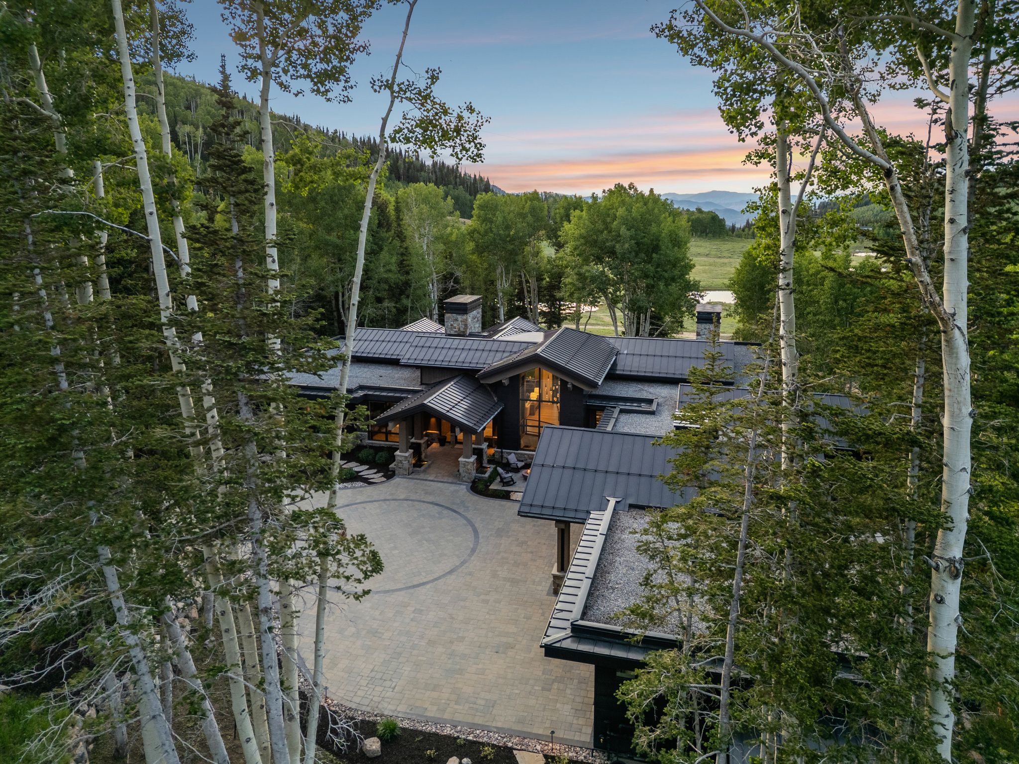 Contemporary mountain residence featuring a multi-gabled metal roof system, stone and wood exterior finishes, a paver circular drive, and a backdrop of mature trees