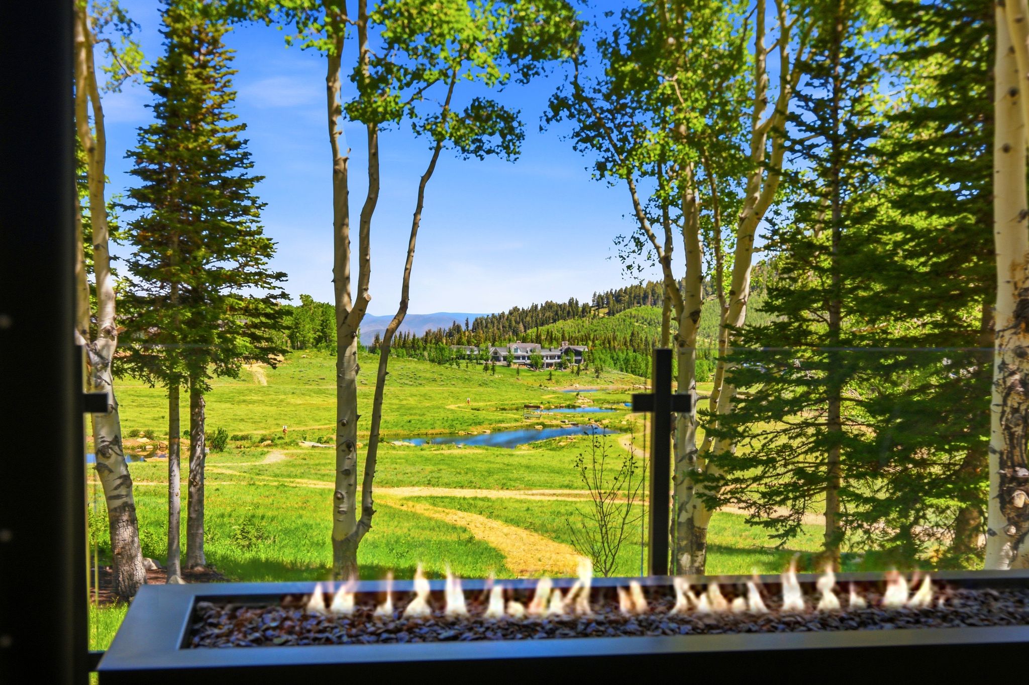 Outdoor fire feature with a stone bed, framed by mature trees, overlooking a verdant meadow with a winding path and distant water features under a clear sky