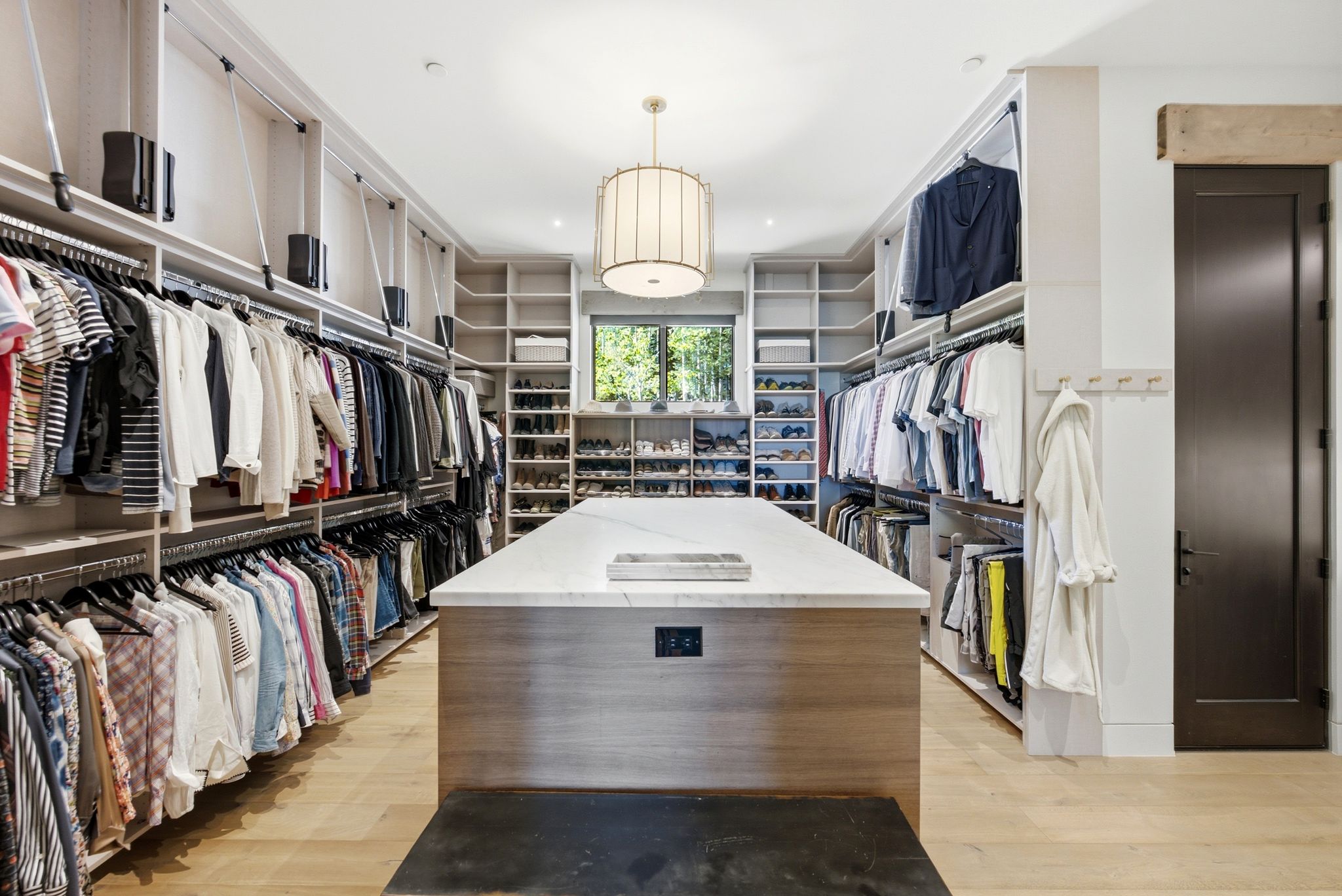 Expansive custom closet featuring extensive built-in shelving, multiple hanging rods, a central island with a light-toned countertop, wood-finish flooring, and a window providing natural illumination