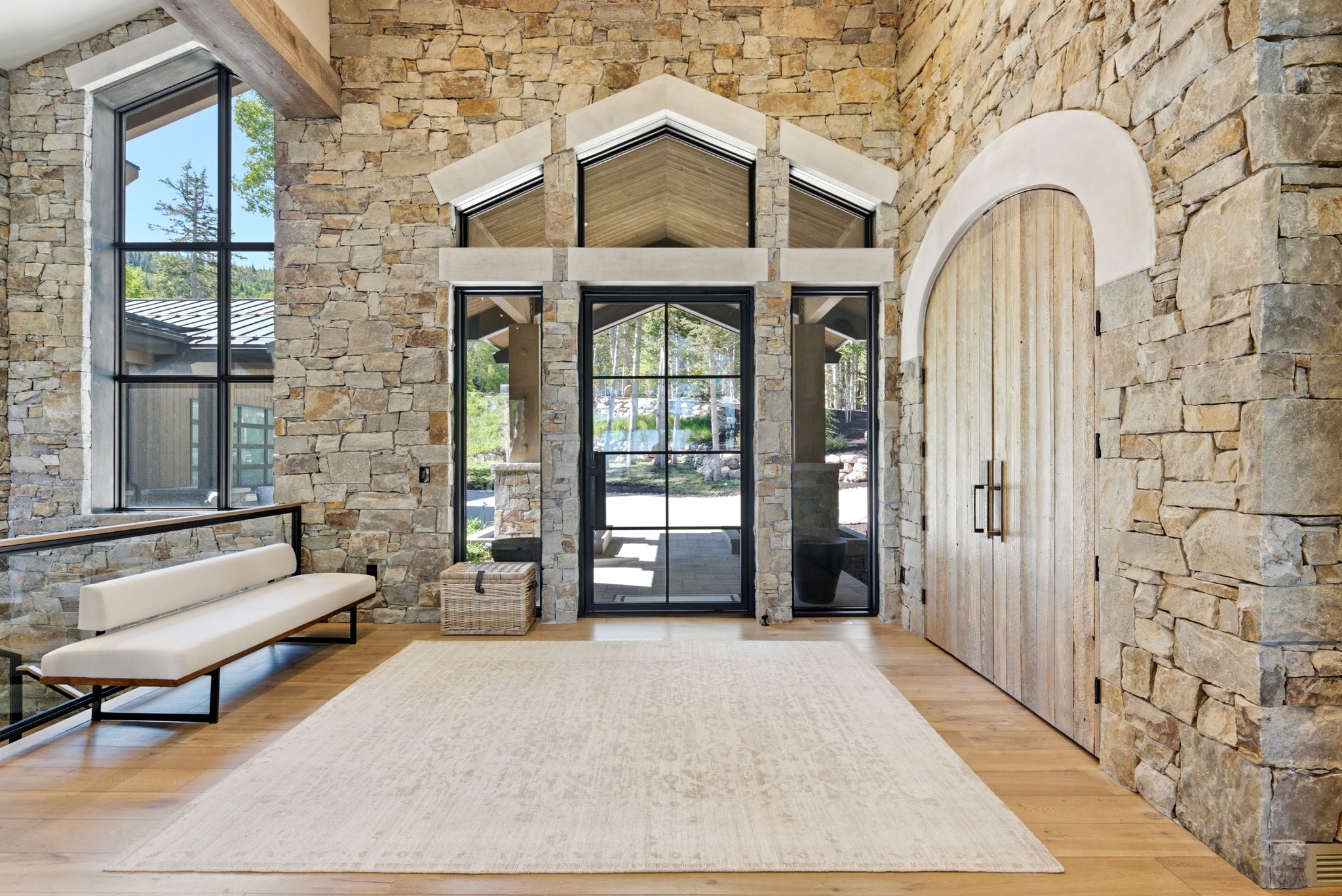 Stone wall entry featuring an arched wooden door, black-framed glass entrance, large vertical window, exposed wood beam ceiling, and light wood-finish flooring