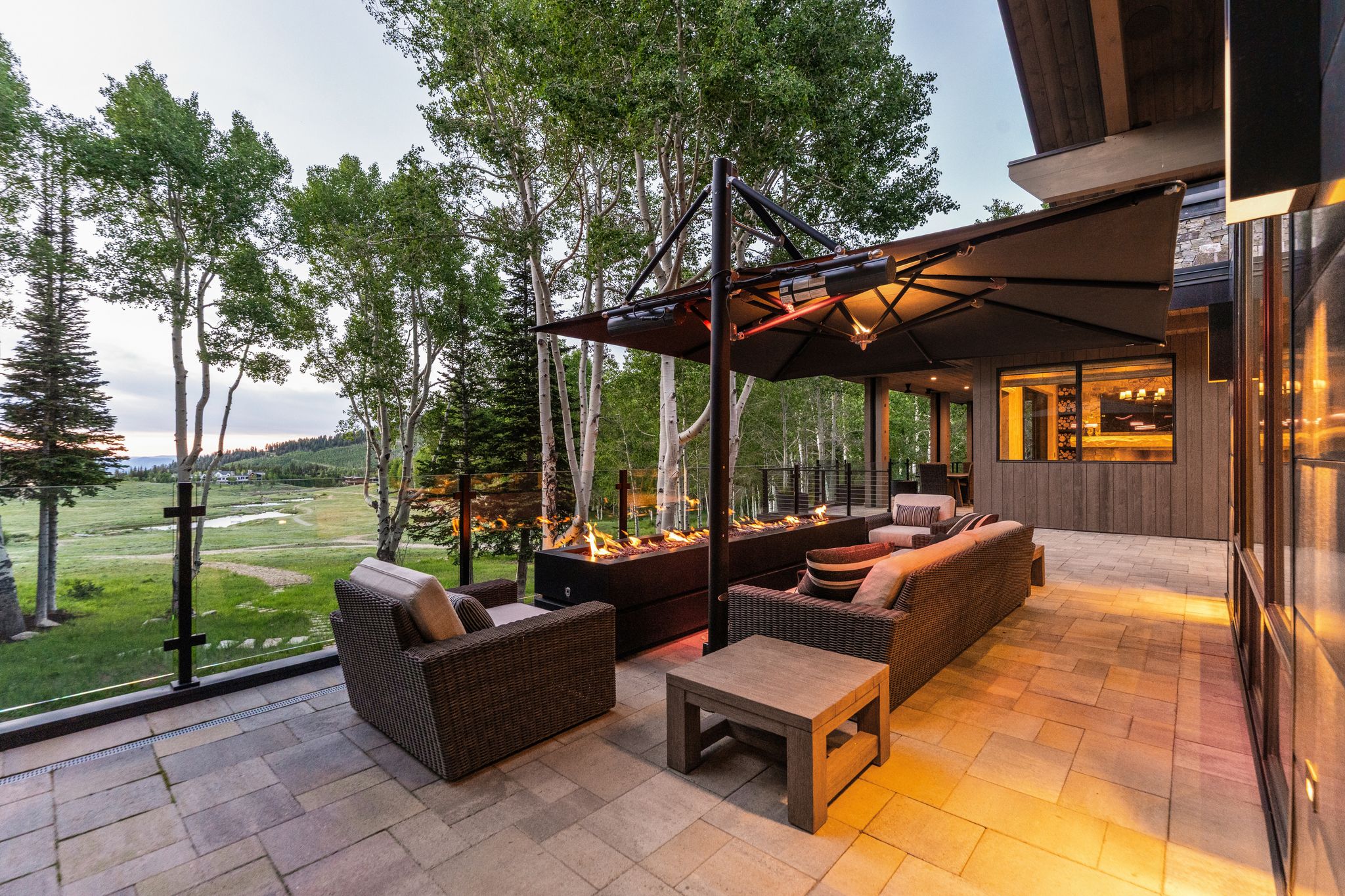 Expansive stone patio featuring a built-in fire pit, glass railing, and a large umbrella with integrated lighting