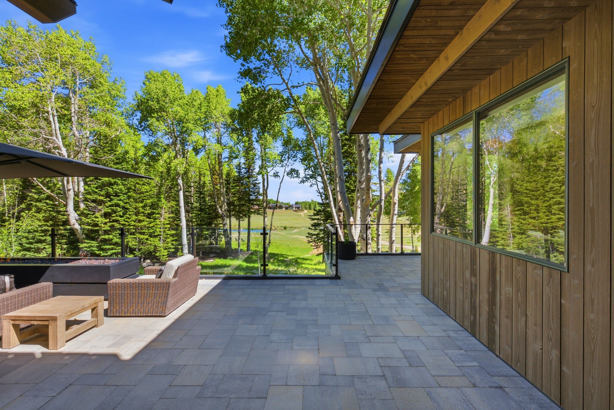Spacious outdoor patio featuring stone paver flooring, a glass panel railing system, and a natural wood-clad exterior