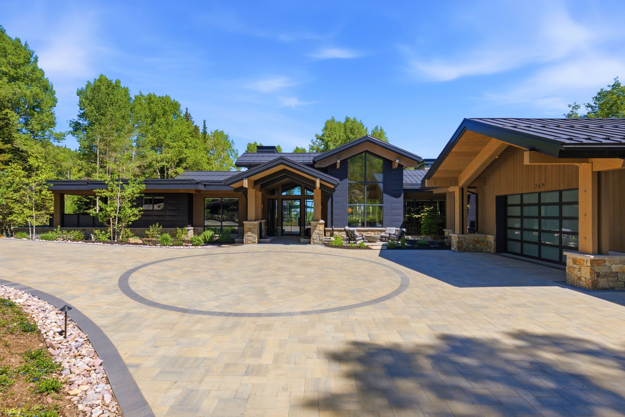 Contemporary mountain-style residence featuring a paved circular driveway with accent banding, a black metal roof, and stone pillars