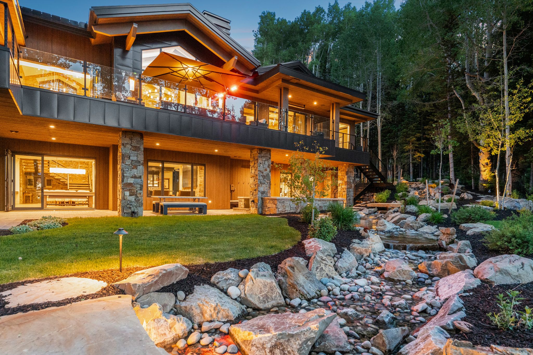 Multi-level property with natural wood siding and stone accents, featuring an upper-level balcony with glass railing, a ground-level patio, and extensive landscaping with a rock-lined water feature