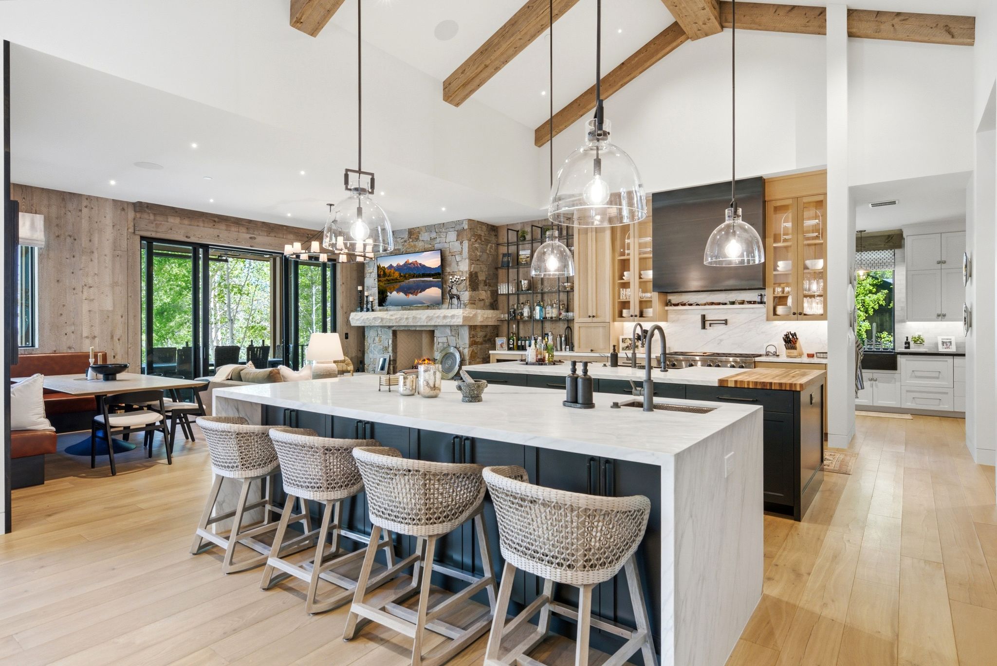 Spacious open-concept kitchen featuring a large waterfall island with seating, natural wood flooring, exposed wood beams, and a stone fireplace