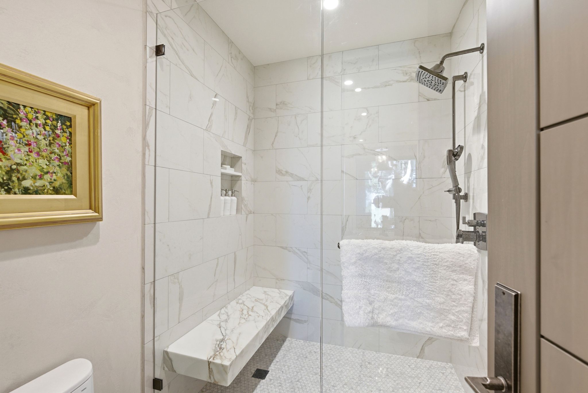 Glass-enclosed shower featuring white stone-look tile walls, a built-in stone bench, a recessed niche, a rain showerhead, and pebble-style flooring