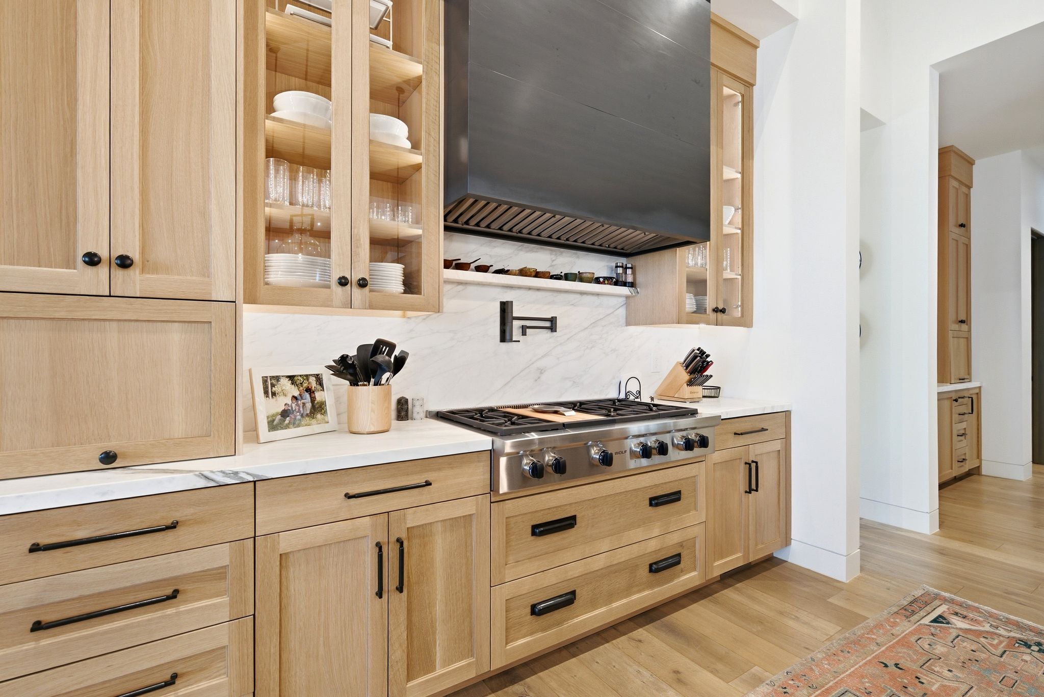 Gourmet kitchen featuring a stainless steel range with a matte black hood, light wood-finish cabinetry, glass-front upper cabinets, white stone countertops, and a white stone backsplash