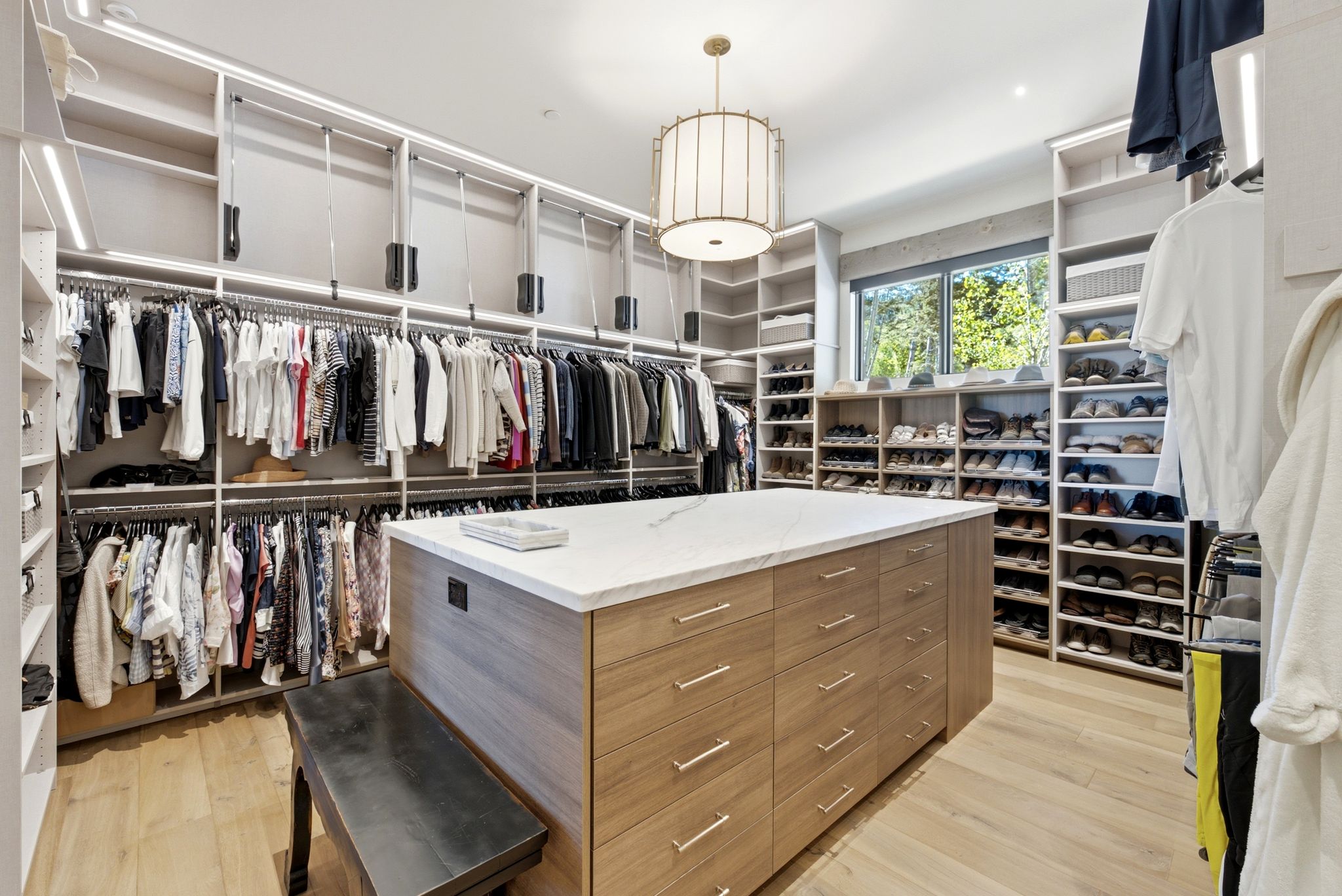 Custom closet featuring extensive built-in shelving, organized hanging storage, a central island with drawers and a light-toned countertop, wood-finish flooring, and a contemporary ceiling fixture