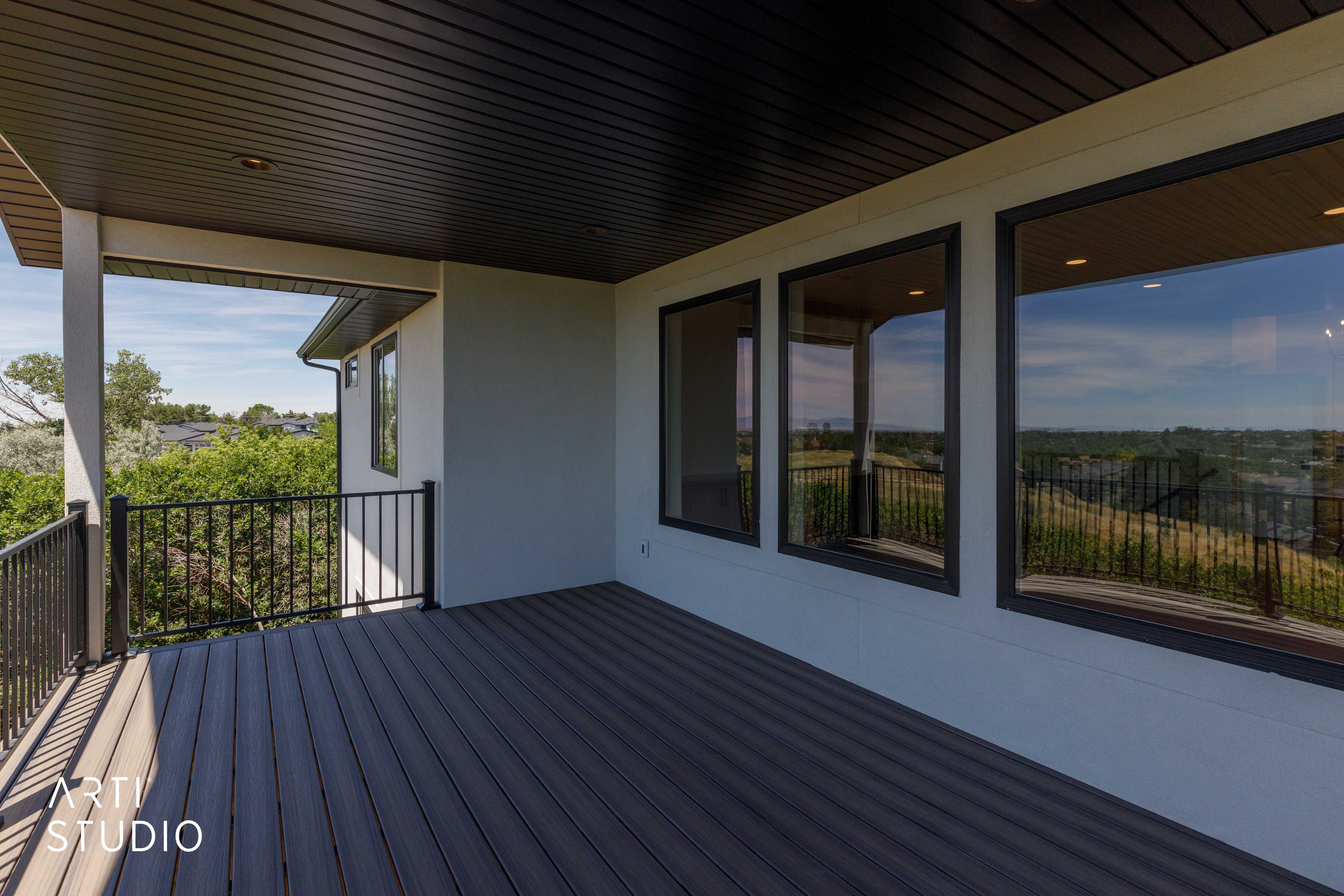 Covered outdoor deck featuring composite decking, black metal railings, recessed lighting, and expansive windows