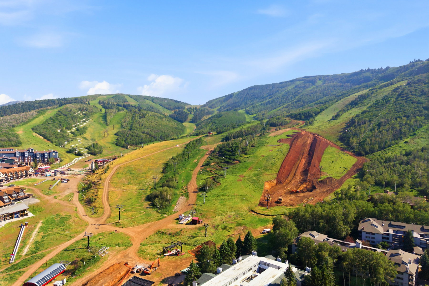 Mountain terrain featuring expansive green slopes, scattered evergreen trees, ski lift structures, and visible development areas
