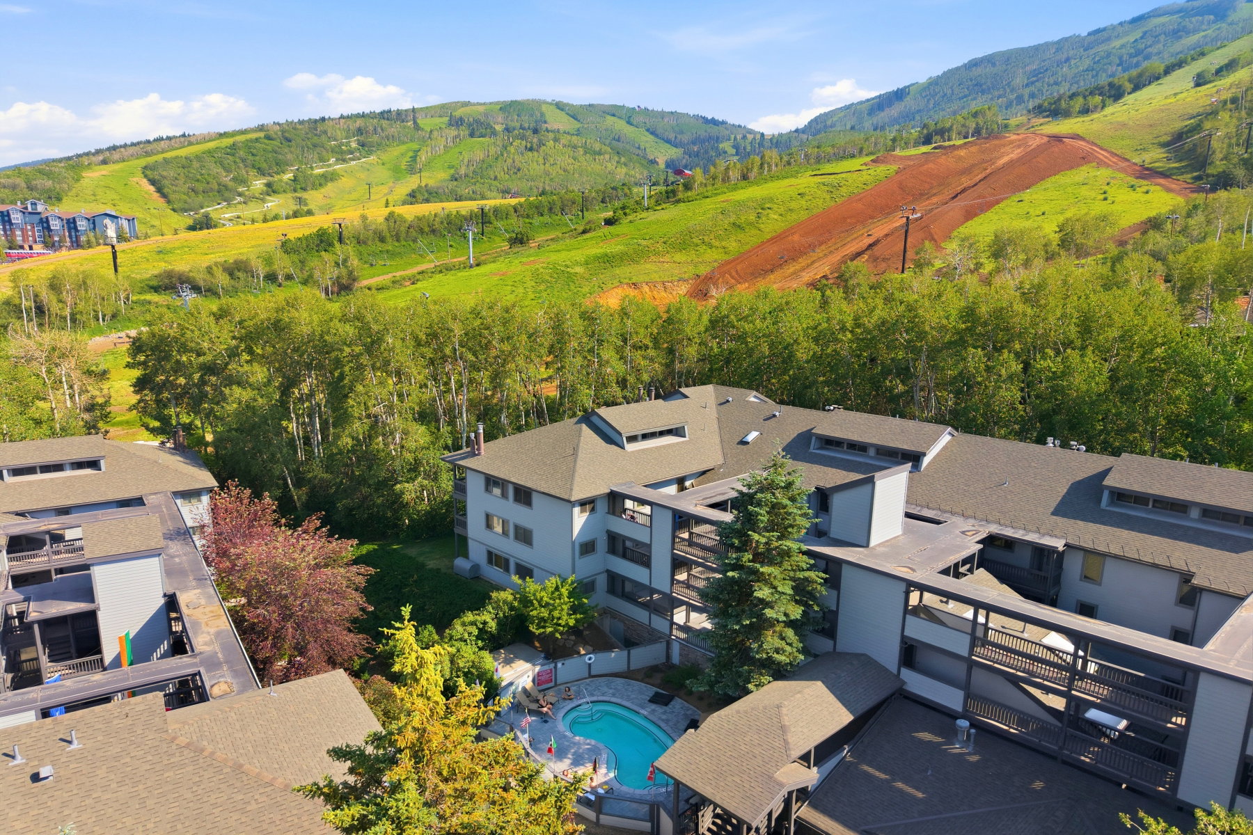 Multi-story residential buildings with balconies, surrounded by mature trees, featuring a central swimming pool and hot tub area