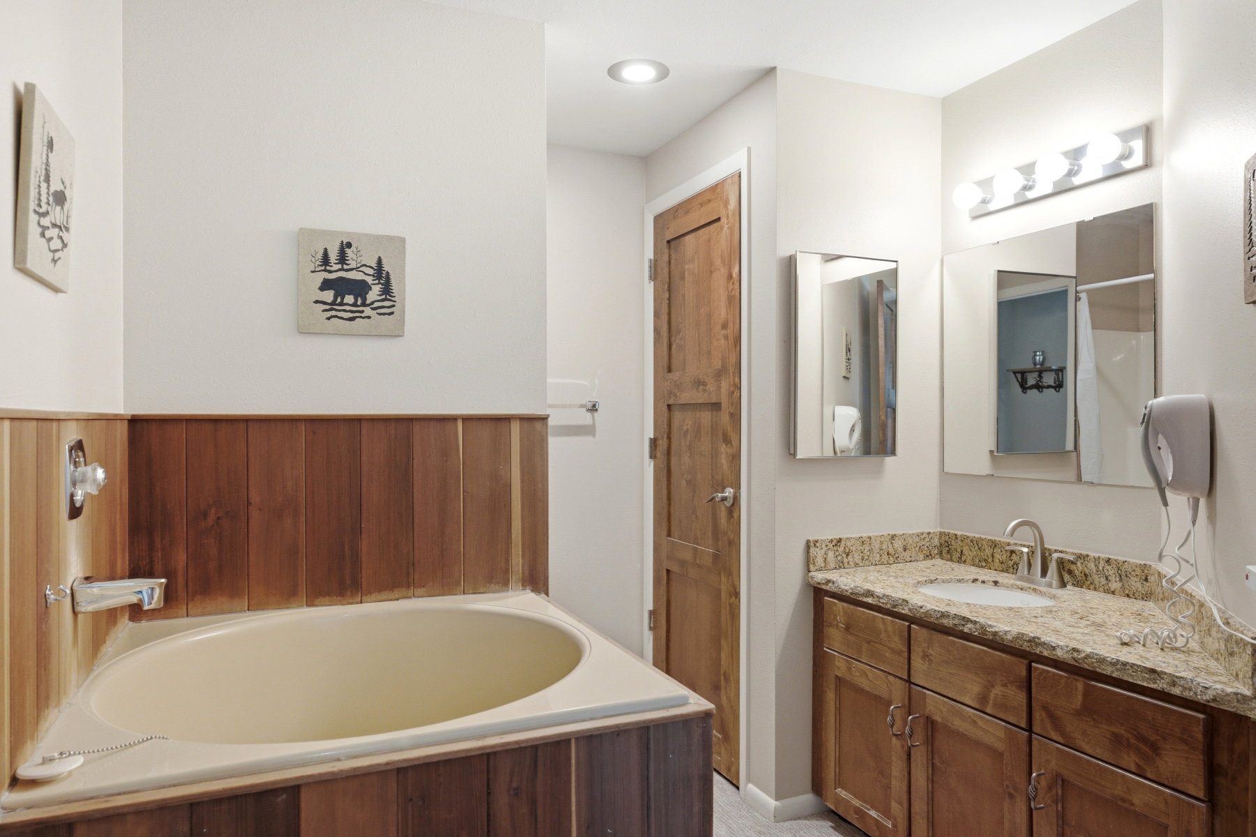 Bathroom featuring a corner soaking tub with wood-panel surround, a vanity with wood cabinetry, a light-toned countertop, and dual mirrors
