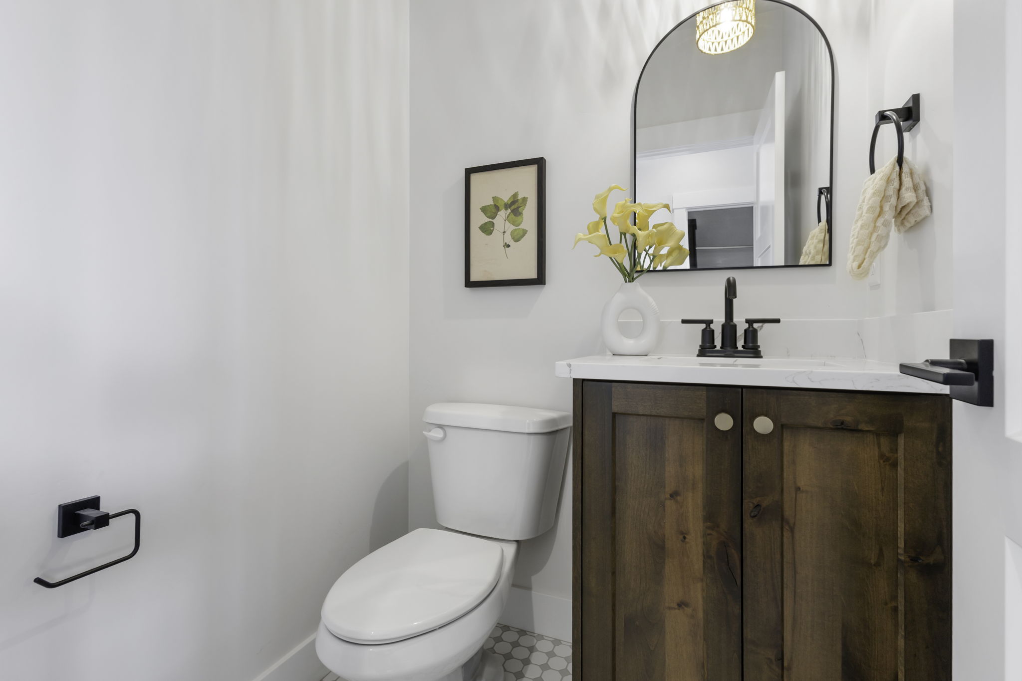 Powder room featuring a wood-finish vanity with a white countertop, an arched mirror, matte black fixtures, and mosaic floor tiling