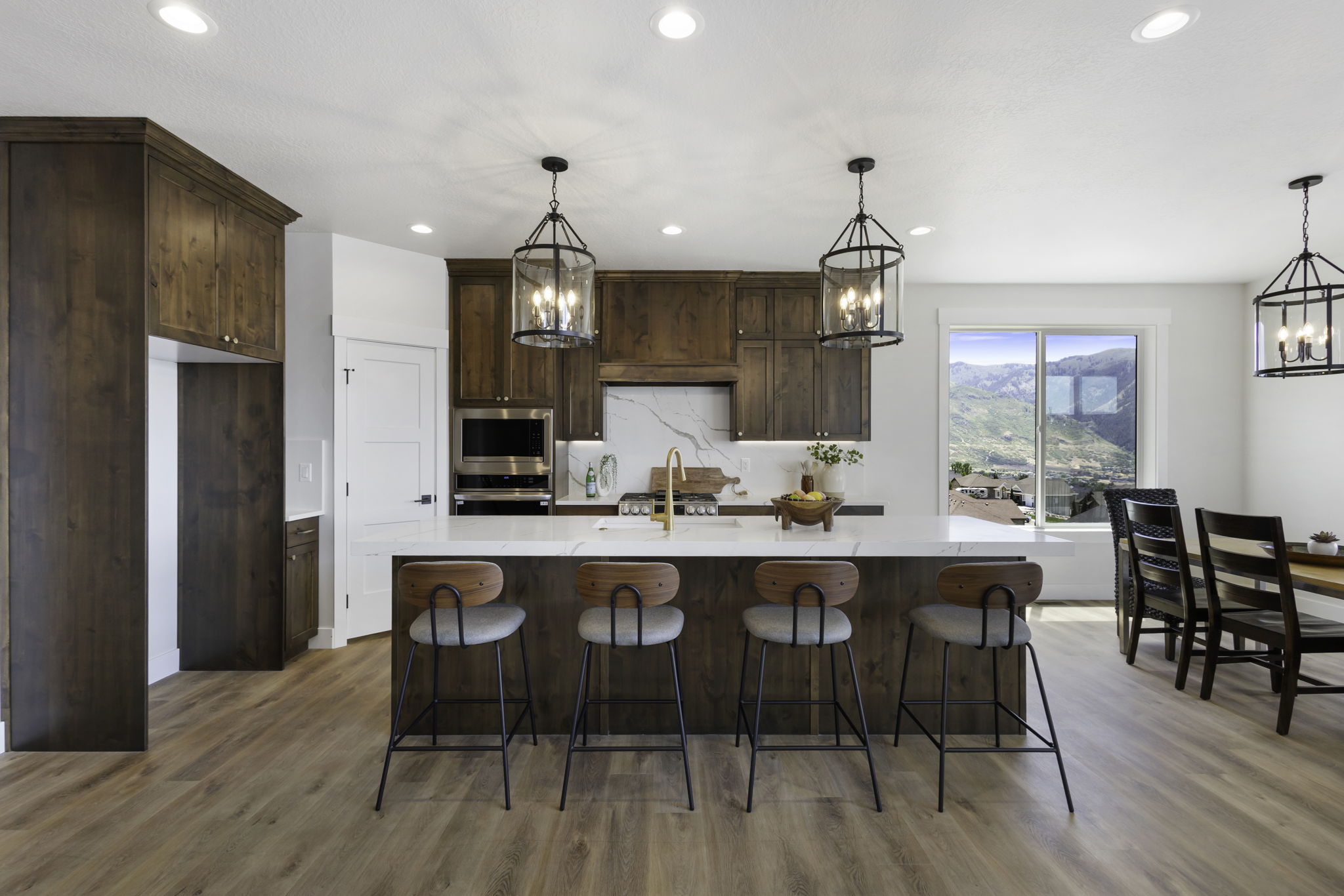 Open-concept kitchen featuring wood-finish flooring, custom dark wood cabinetry, a large kitchen island with counter seating, stainless steel appliances, and a window framing distant mountains
