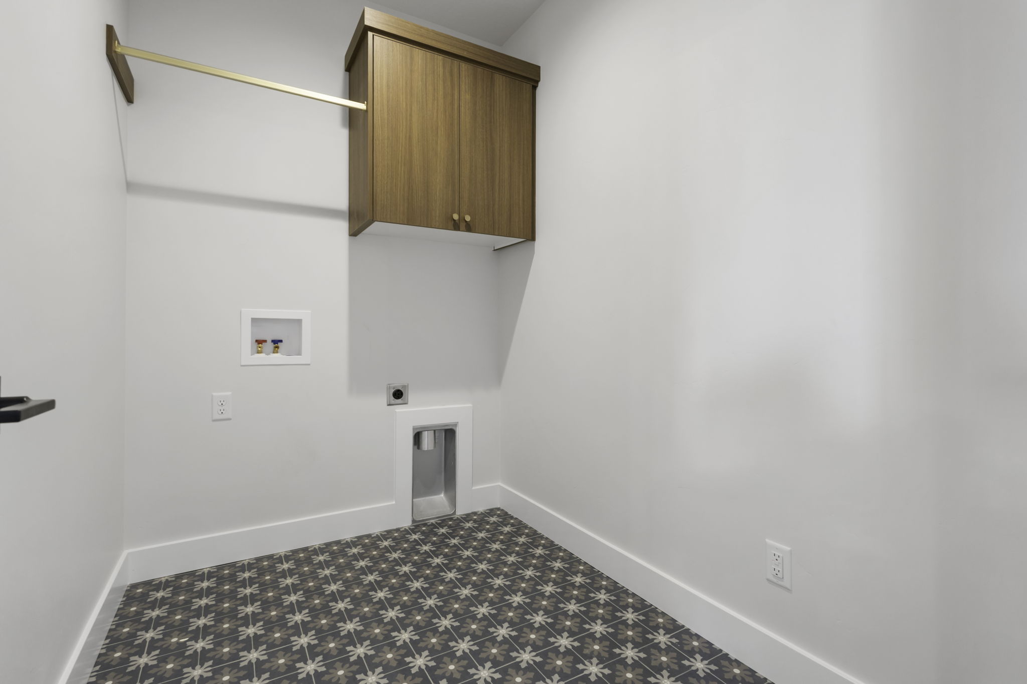 Laundry area featuring patterned floor tiles, a built-in wood-finish cabinet, gold-tone hanging rod, and white baseboards
