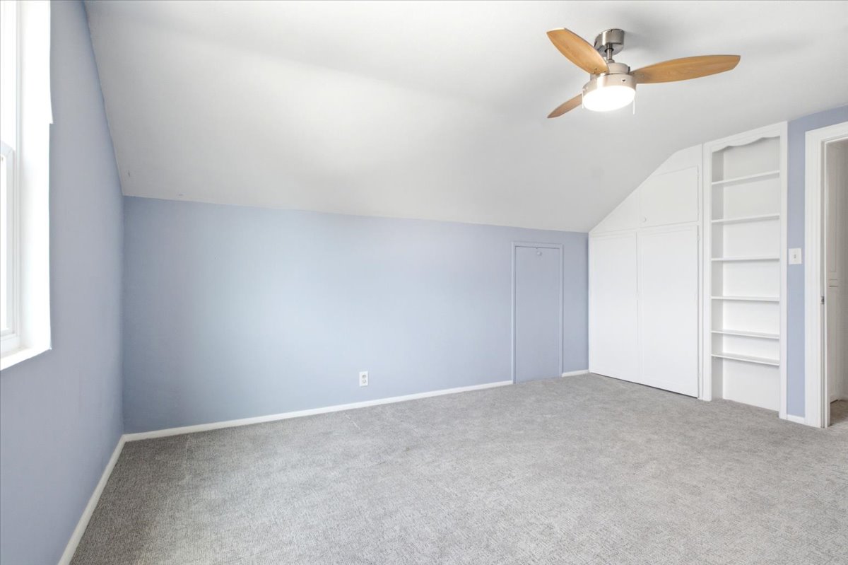 Carpeted room with a vaulted ceiling, a window, a ceiling fan with light fixture, built-in shelving, and a built-in closet with bifold doors