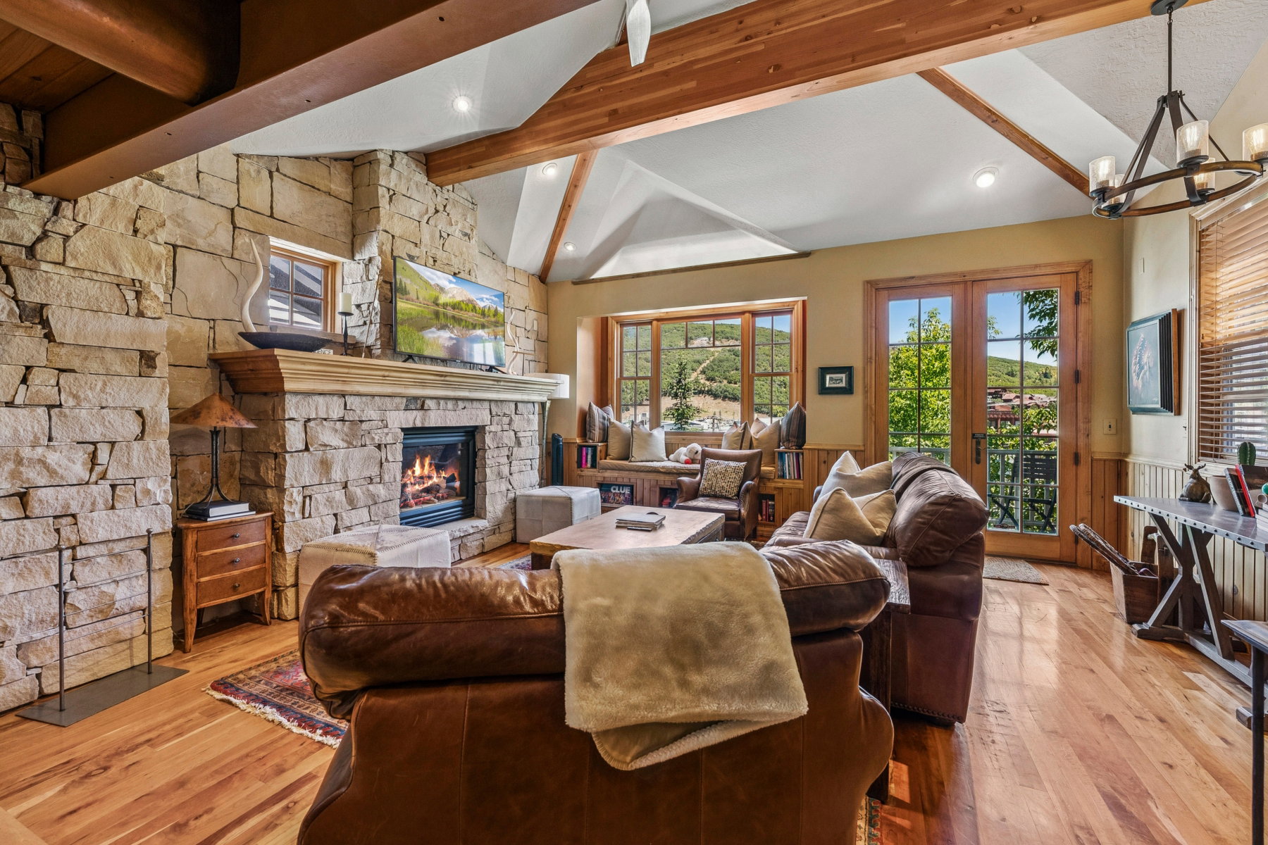 Living area featuring a stone fireplace with a wood mantle, wood-finish flooring, vaulted ceilings with exposed wood beams, and French doors opening to an exterior setting