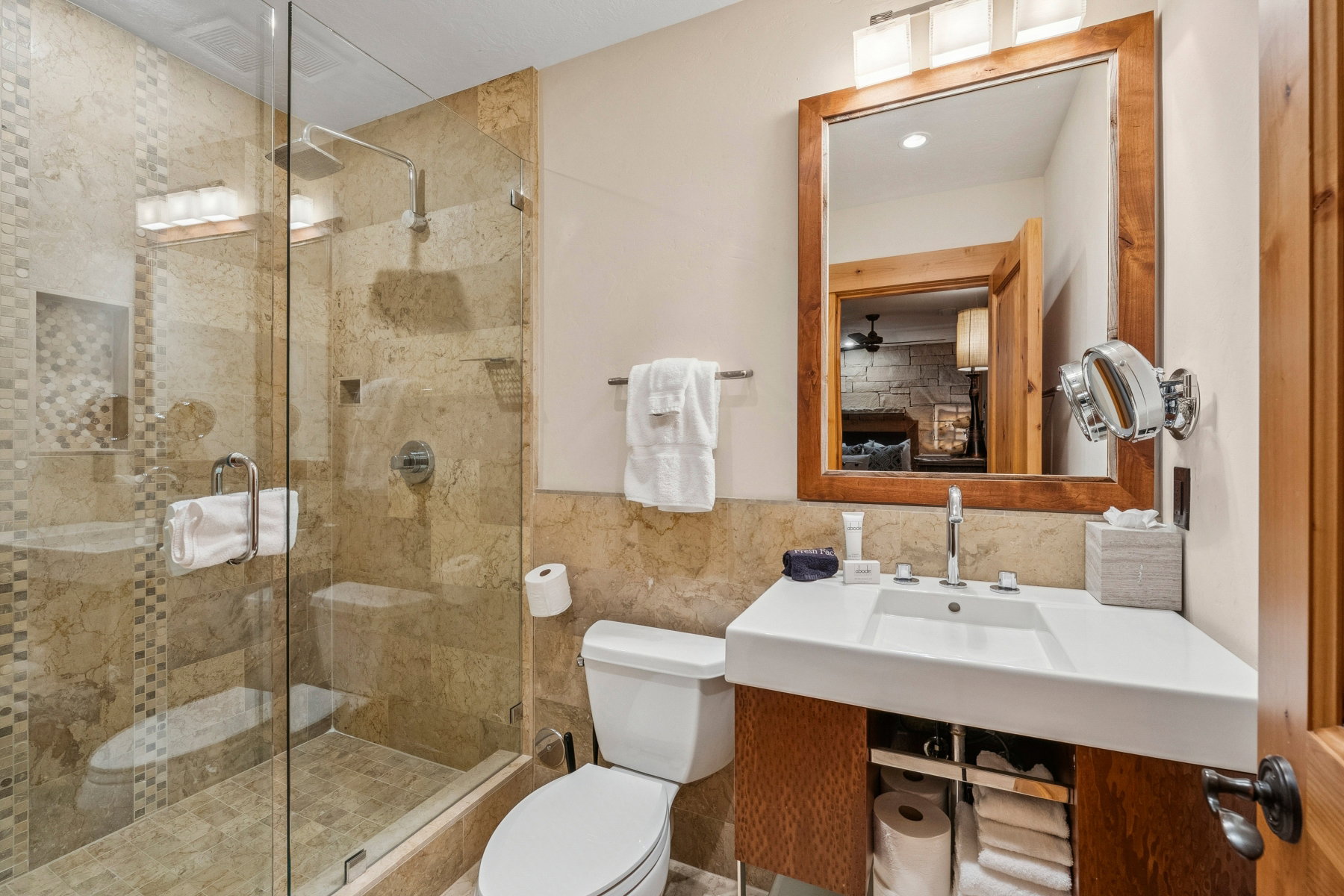 Full bathroom featuring a glass-enclosed shower with a rainfall showerhead, stone-tiled walls, a white vanity with integrated sink, and an unframed wood-finish mirror
