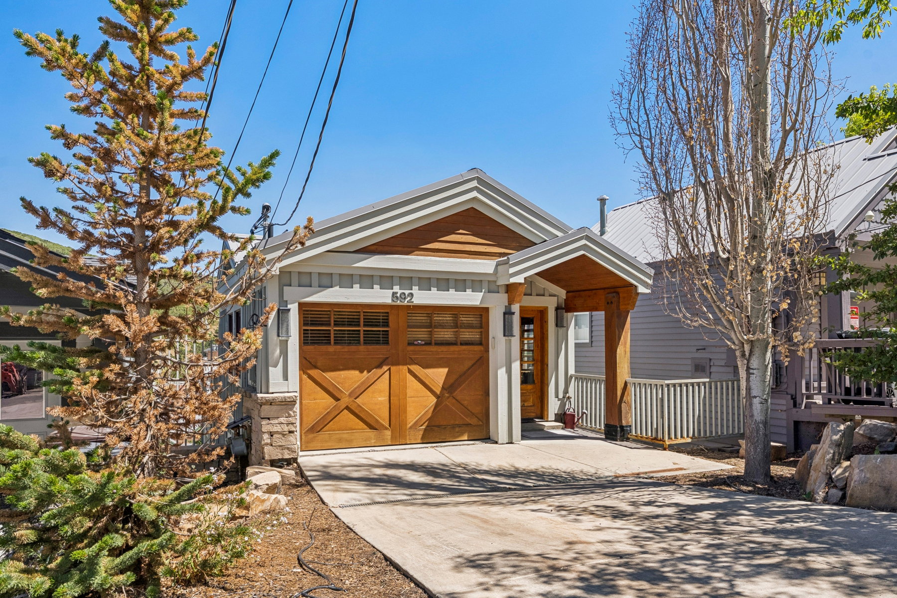 Contemporary single-story residence featuring a wood-panel garage door with decorative cross-bracing, a covered front porch with a wood-clad ceiling, light gray siding, and a metal roof