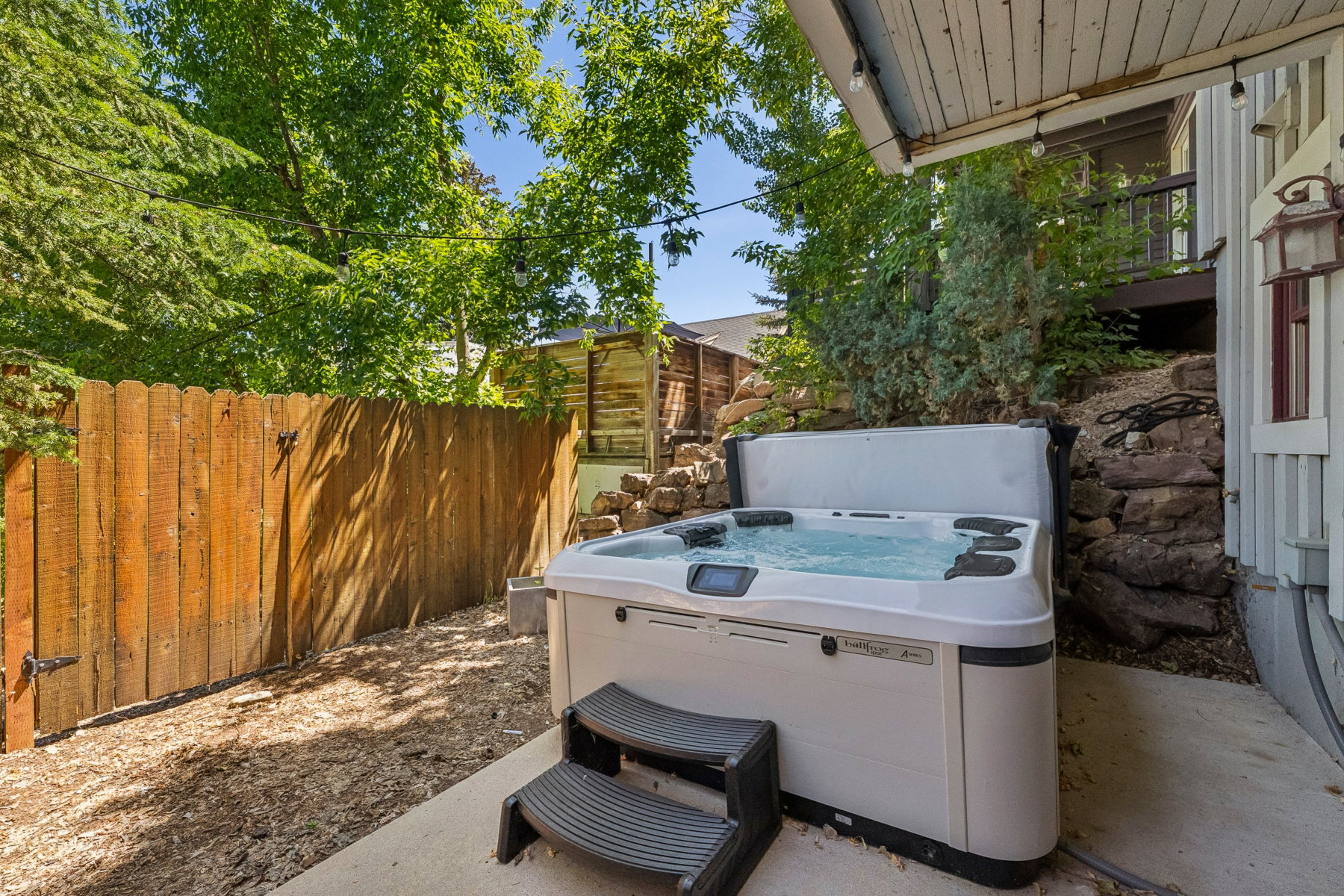 Outdoor spa with steps on a concrete pad, surrounded by natural wood fencing and mature trees