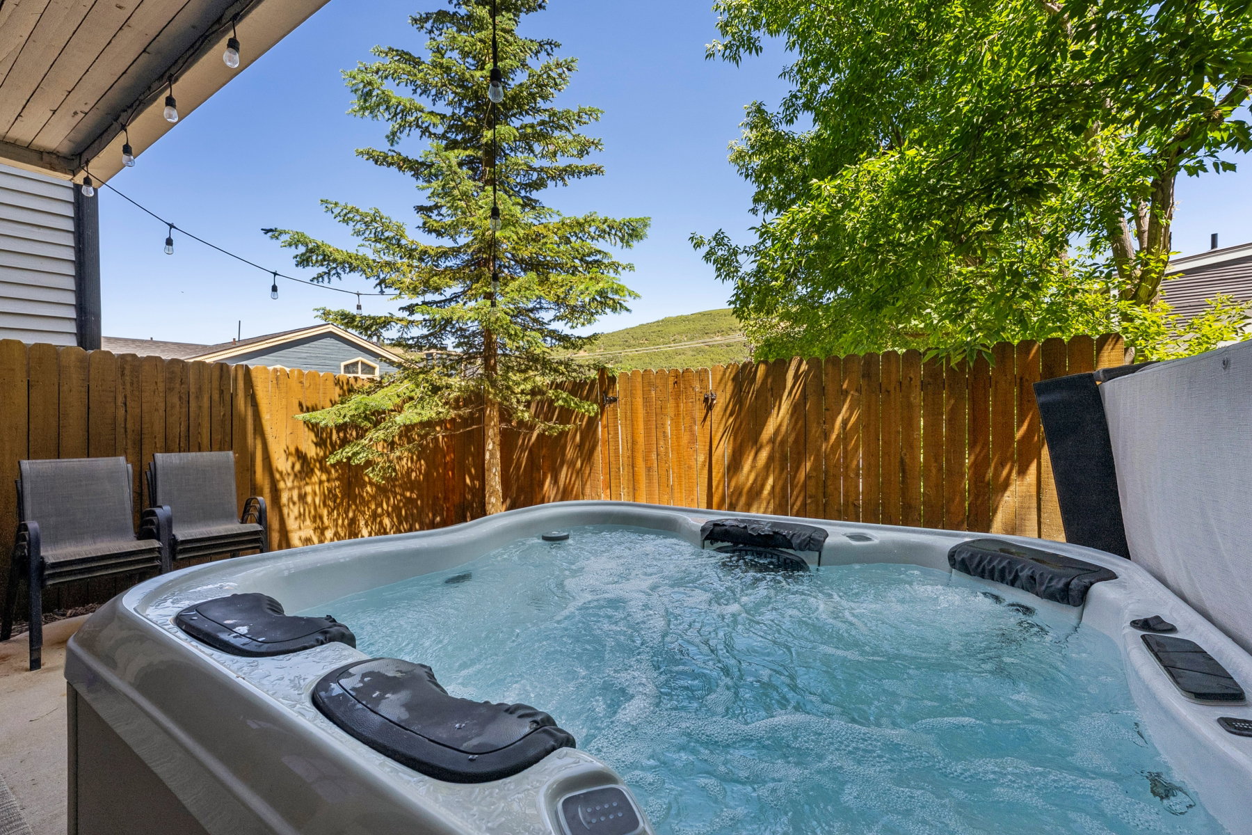 Outdoor hot tub with flowing water, featuring a concrete patio, wood privacy fencing, string lighting, and mature trees