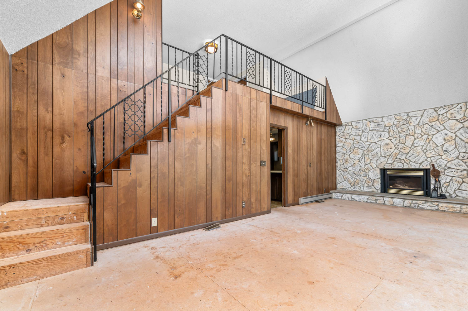 Interior featuring a prominent stone fireplace with a hearth, wood paneling throughout, an open staircase with wrought iron railing, and a loft area