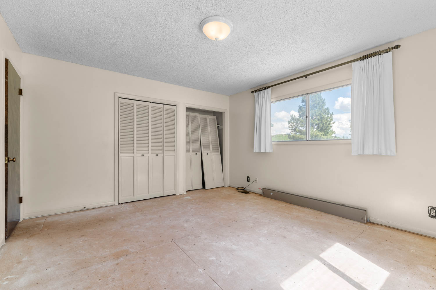 Neutral-toned interior featuring a wide window, two bi-fold louvered closet doors, a flush-mount ceiling light fixture, and textured ceilings