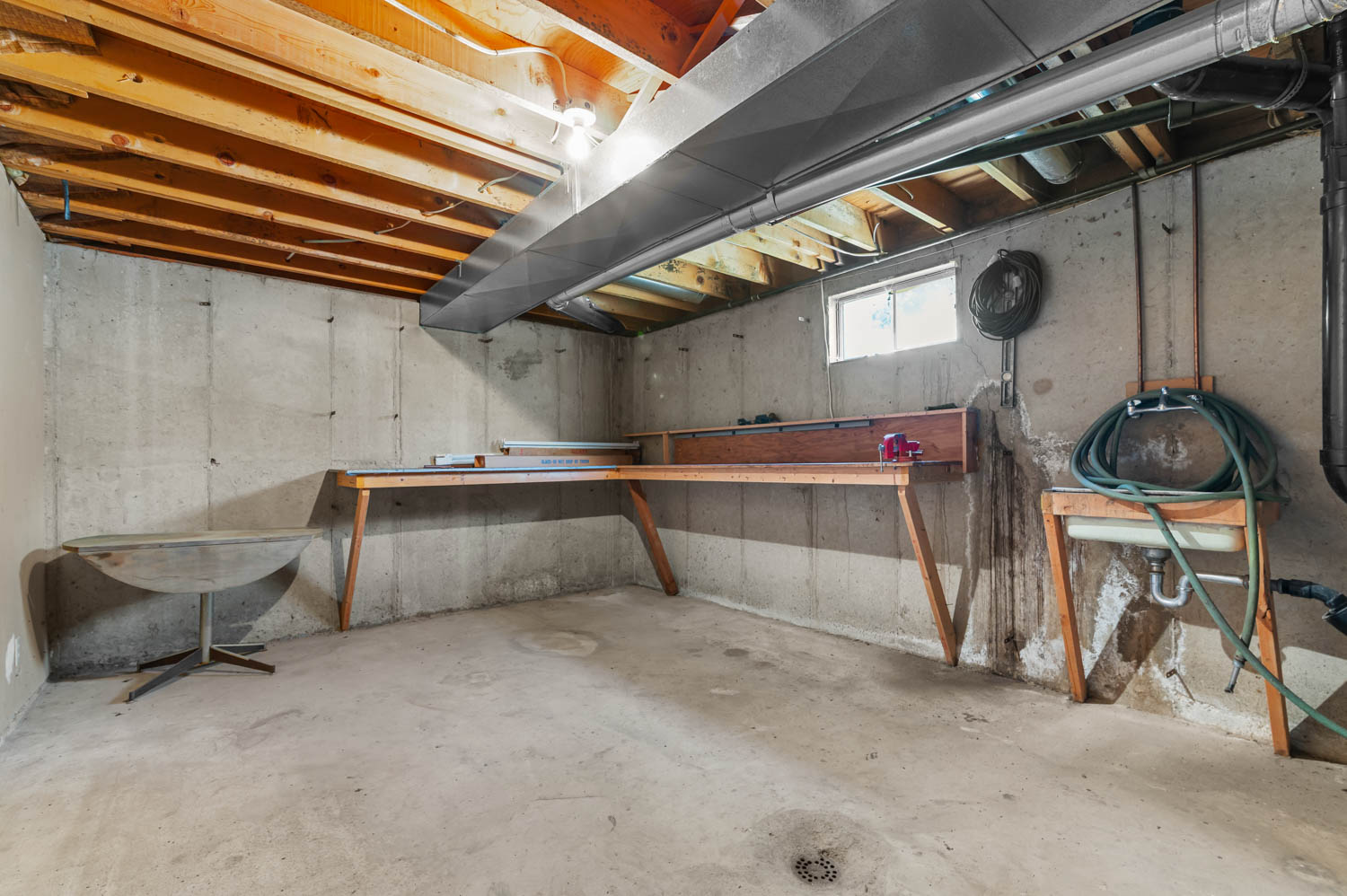 Unfinished basement featuring exposed wood ceiling joists, concrete walls, concrete flooring, a utility sink, and a built-in workbench