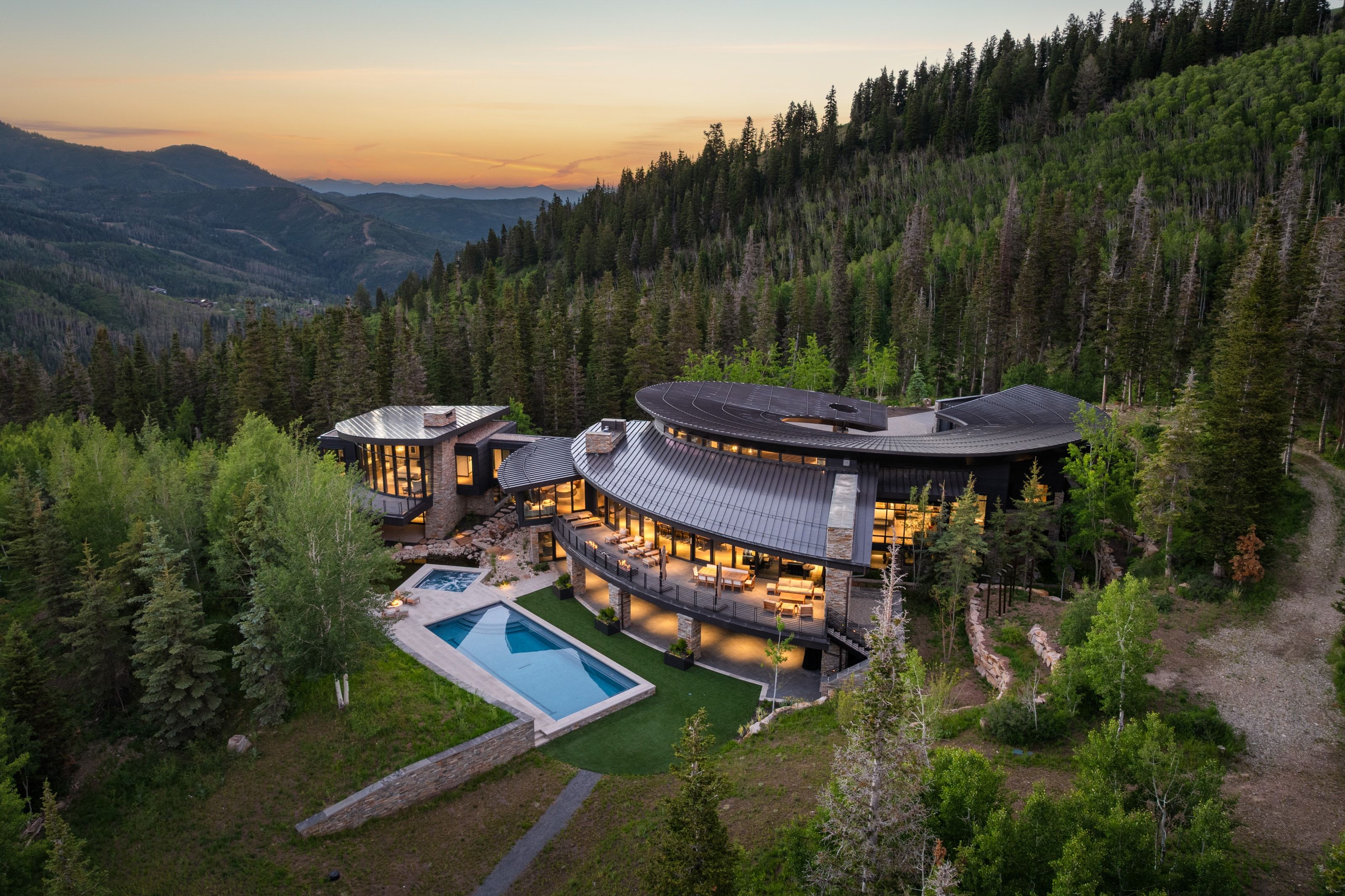 Contemporary mountain residence featuring a curved architectural design, extensive glazing, multiple outdoor terraces, an outdoor swimming pool, and an outdoor spa, all surrounded by dense forest