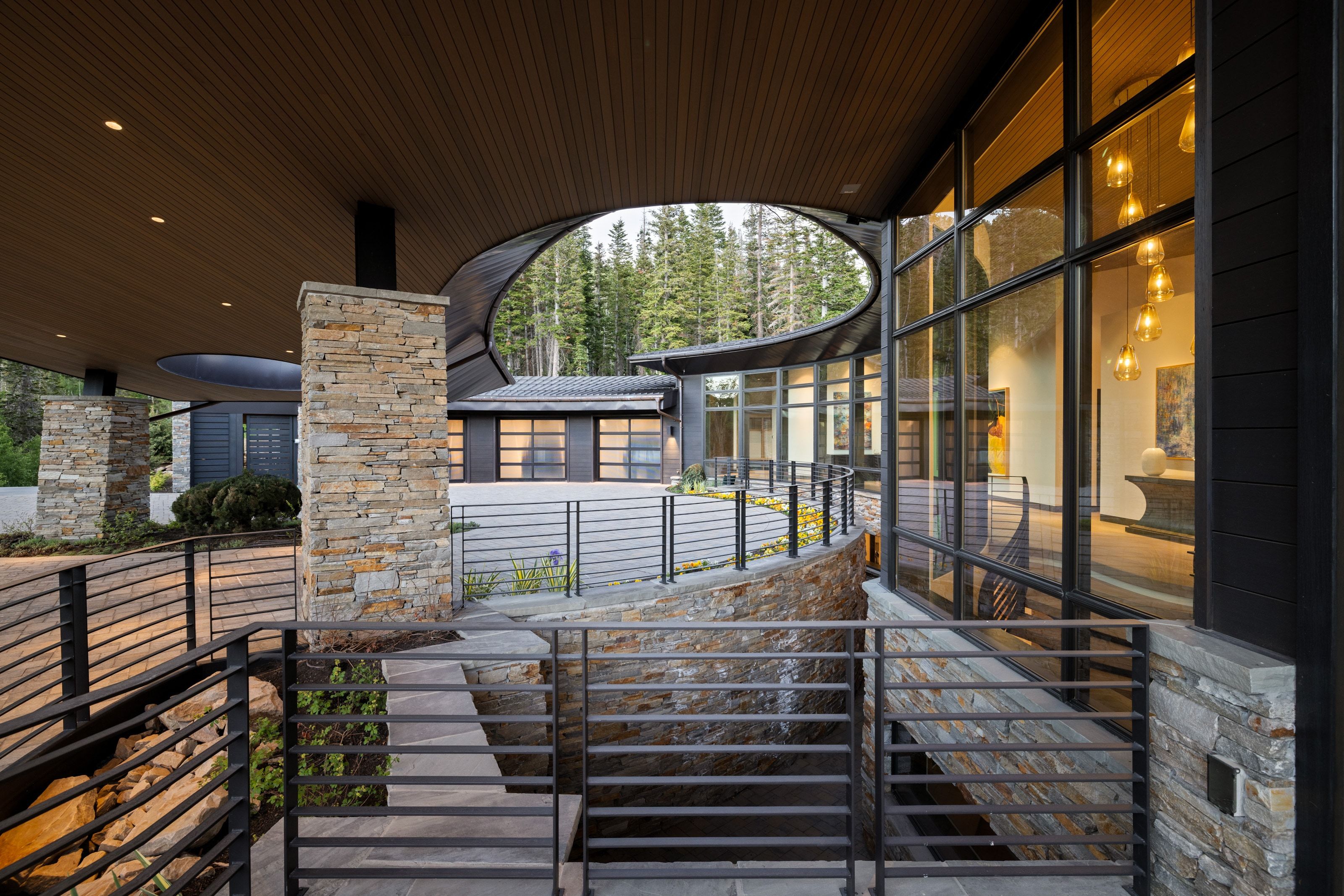 Architectural exterior featuring a curved overhanging roofline with recessed lighting, floor-to-ceiling windows, stacked stone columns, horizontal metal railings, and a forested backdrop