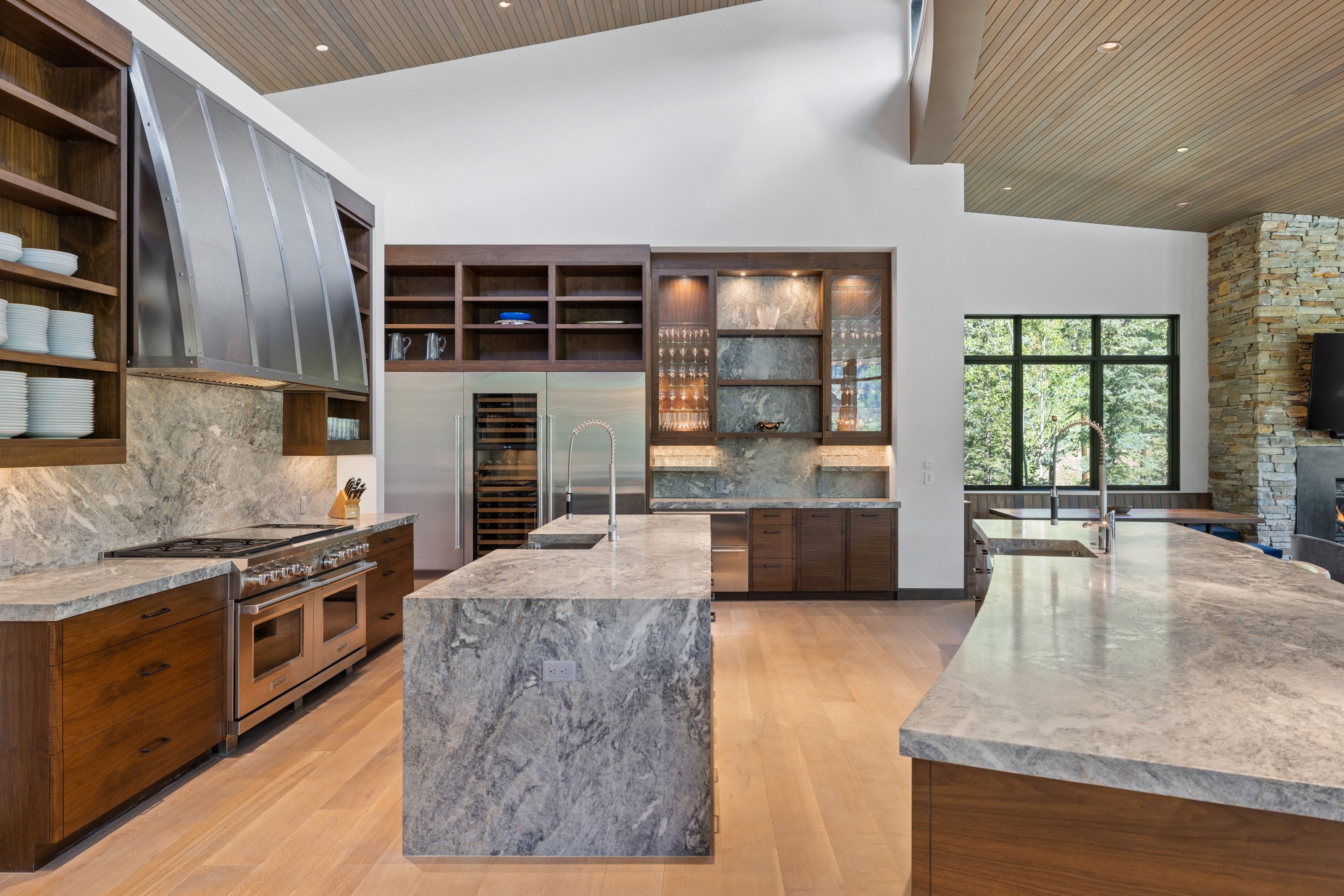 Expansive kitchen featuring a large stainless steel range, dual islands with undermount sinks, wood-finish flooring, integrated wine refrigerator, and vaulted ceilings with wood paneling