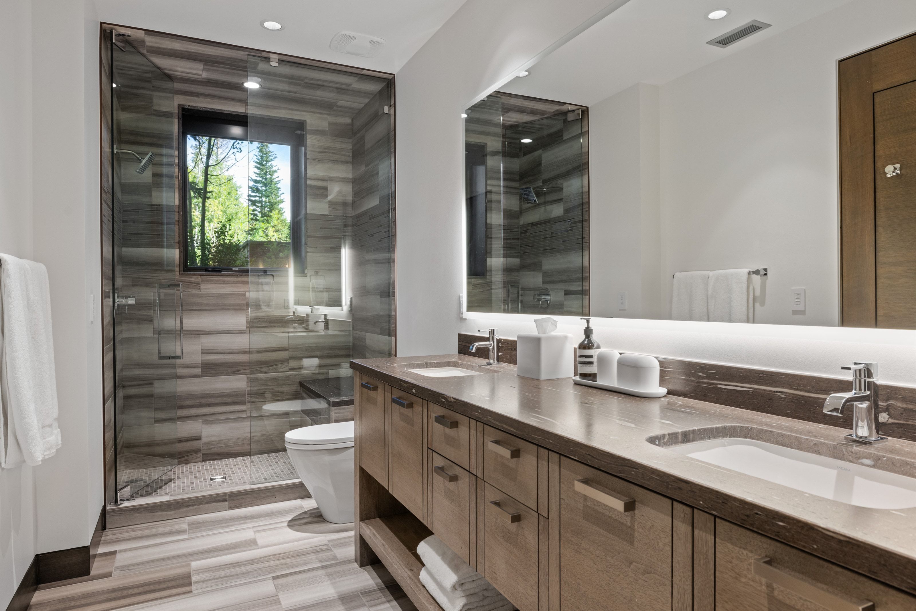Modern bathroom featuring a dual vanity with undermount sinks, a large back-lit mirror, a glass-enclosed shower with a built-in bench, and wood-finish tile flooring